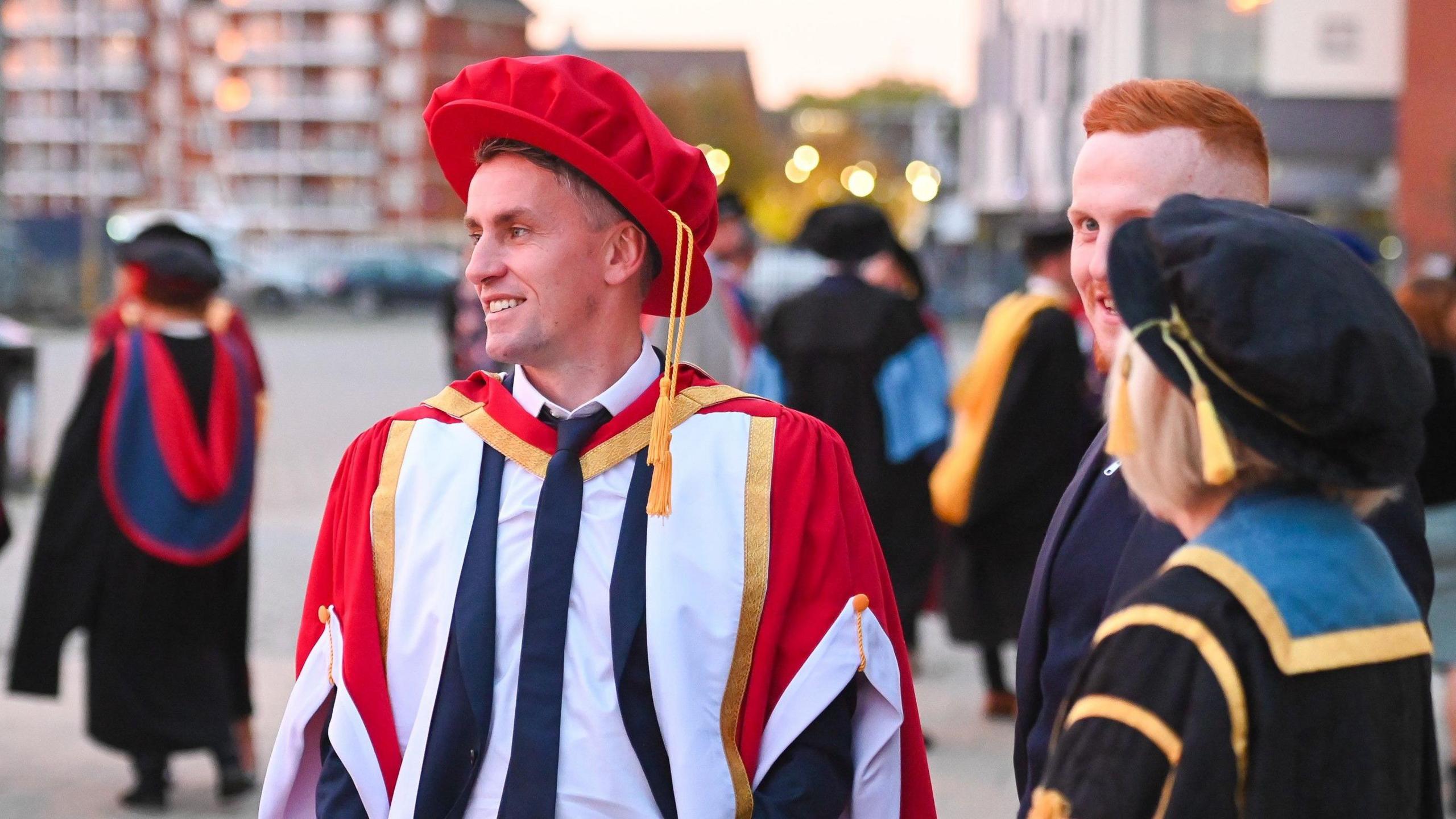 Kieran McKenna wearing a white shirt, dark suit and dark tie plus a red and yellow academic robe and a red academic hat. He is outside and other people wearing academic robes are standing nearby. 
