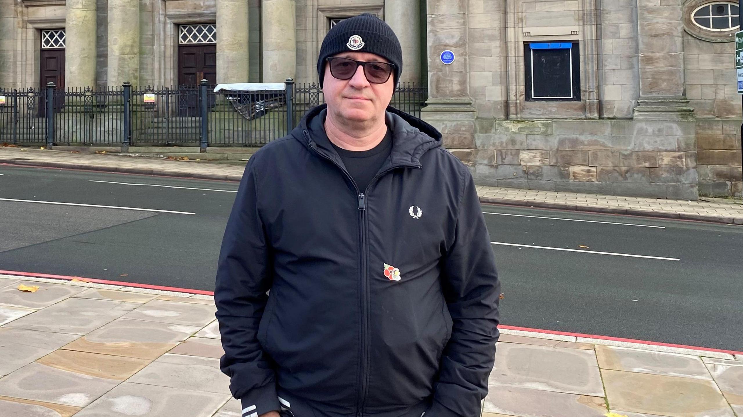 A man wearing a black beanie, sunglasses and a black coat, stands with his hands in his pocket. He is standing on a pavement and behind him across the road is an old, large building.