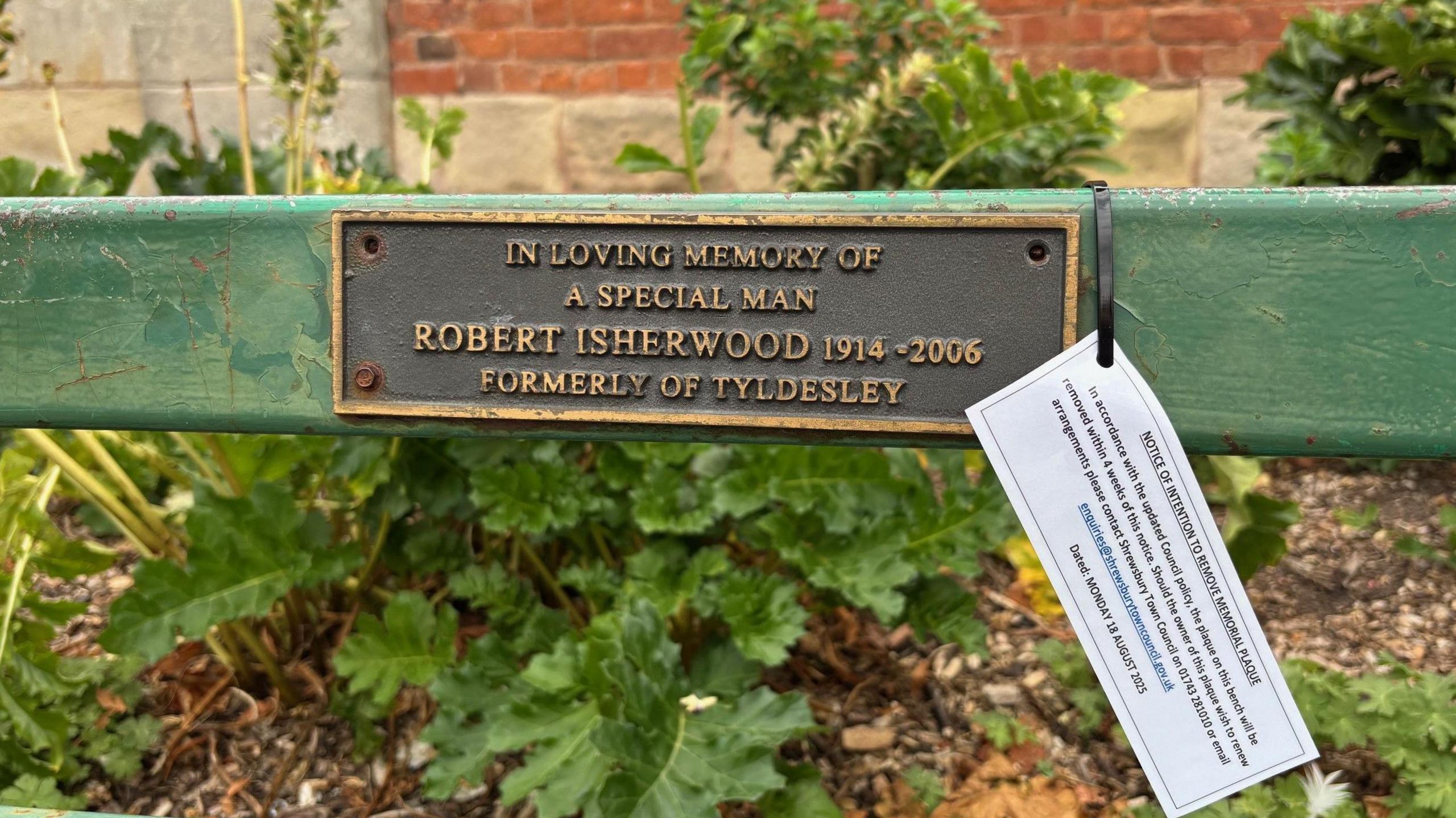 A white sign paper sign, hangs from a bronze-coloured plaque attached to a green park bench. There is greenery behind the bench in a bed, with a wall behind it.    