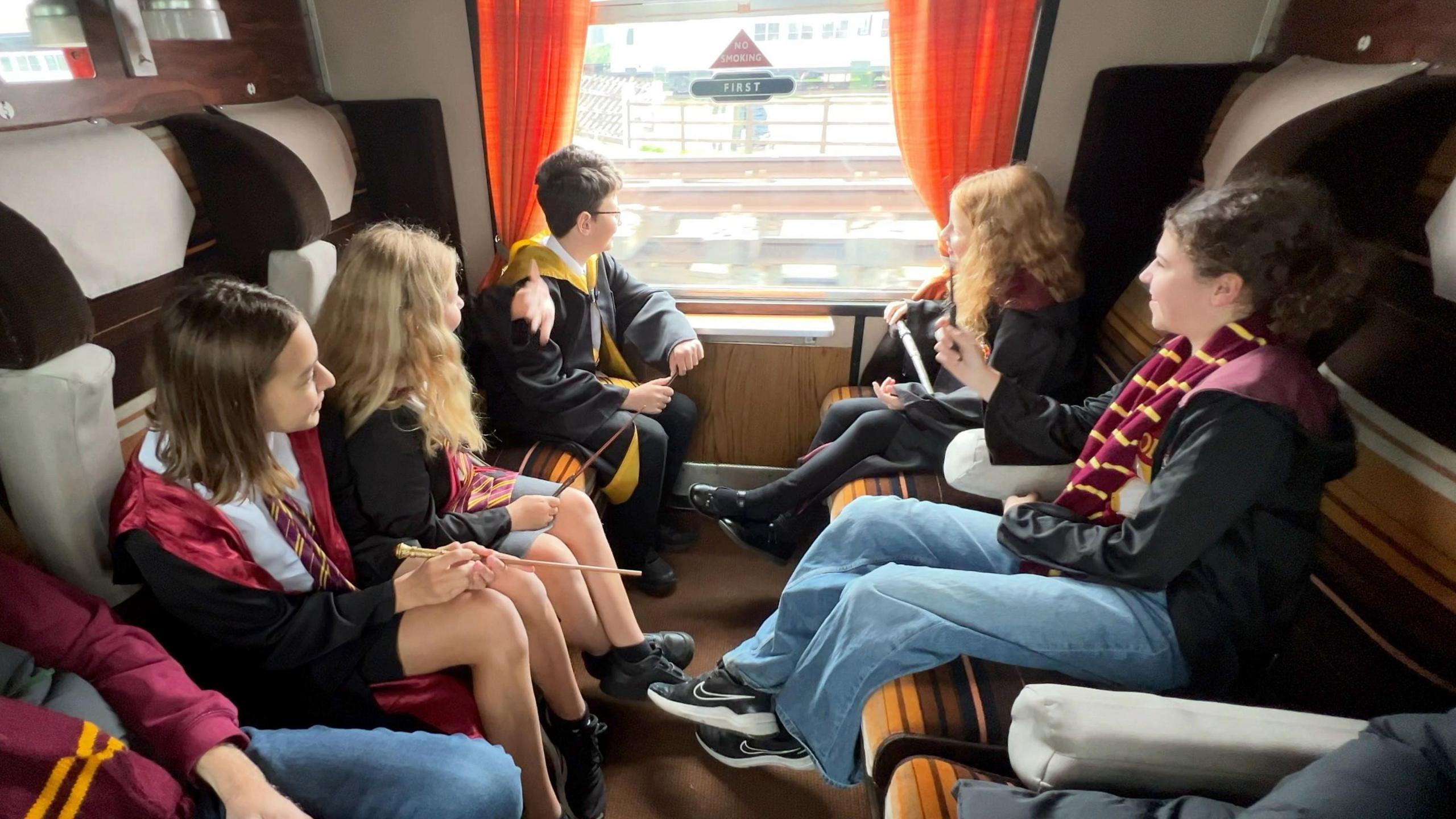 Children carrying wands and wearing uniforms for Hogwarts school, inside one of the compartments on a carriage pulled by the locomotive. They are all looking eagerly out of the window, which has orange curtains.