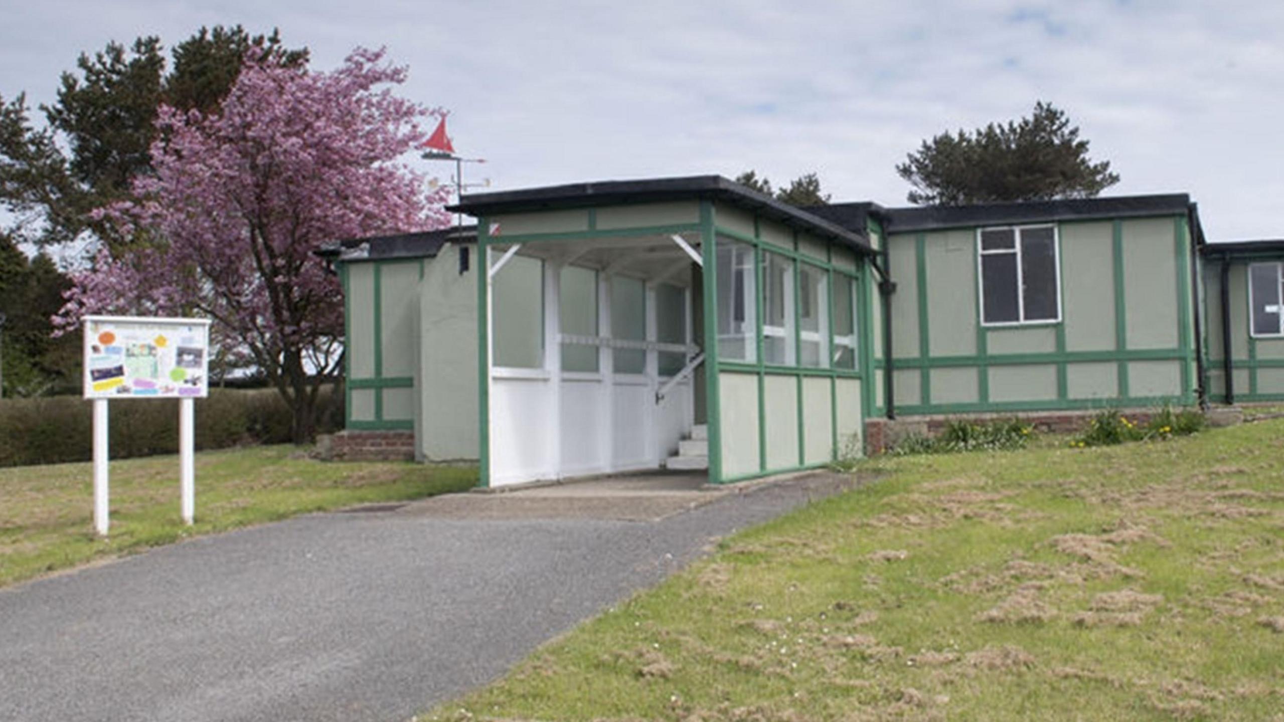 A single-level white building with a black roof and green wooden beams. The sky is overcast and the centre is surrounded by grass.