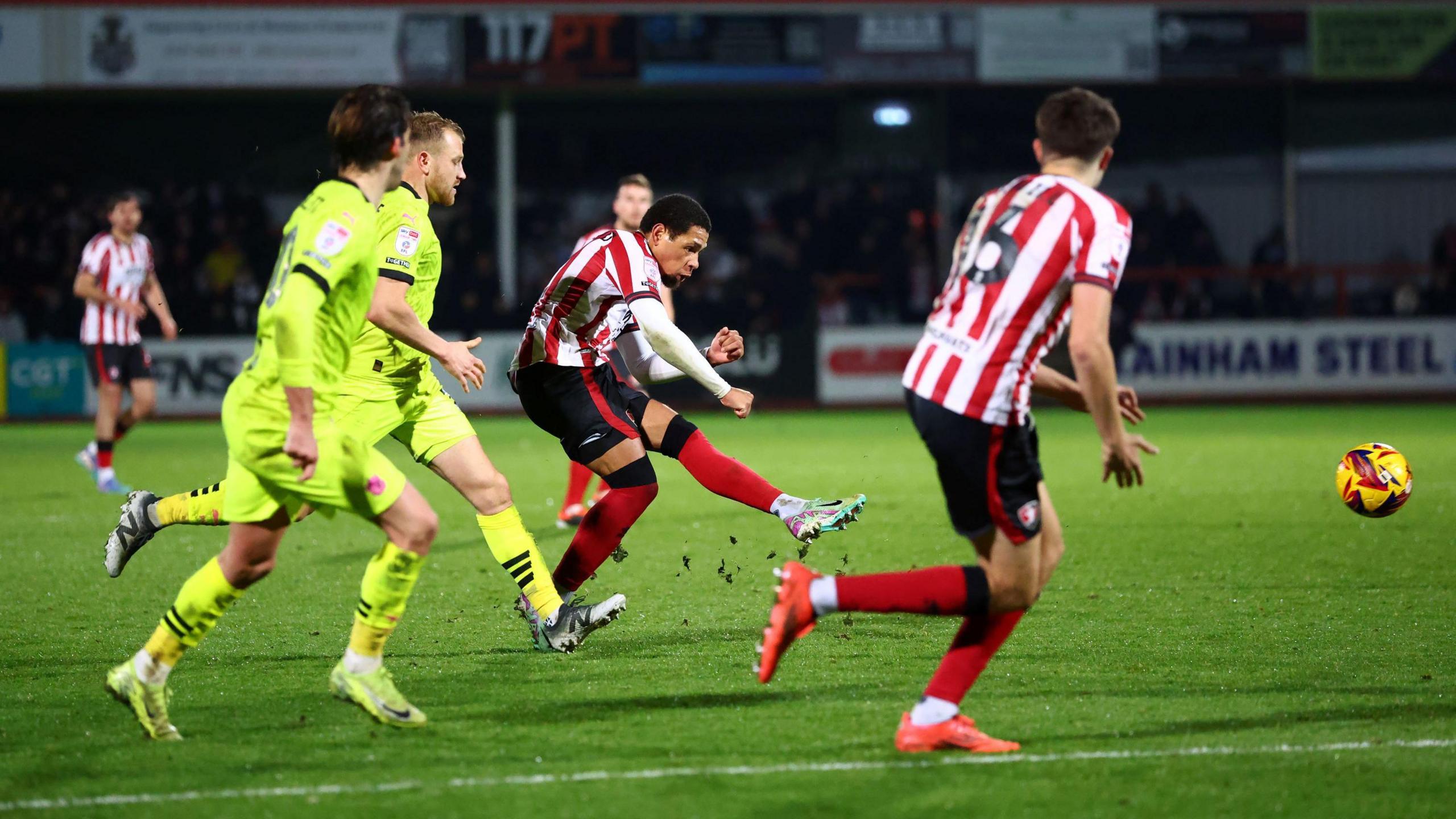 Arkell Jude-Boyd of Cheltenham Town shoots at goal during the match with Port Vale. He is wearing the traditional red and white striped kit, while nearby Port Vale players are wearing their all-yellow change kit. The match is being played at Whaddon Road in Cheltenham under the floodlights