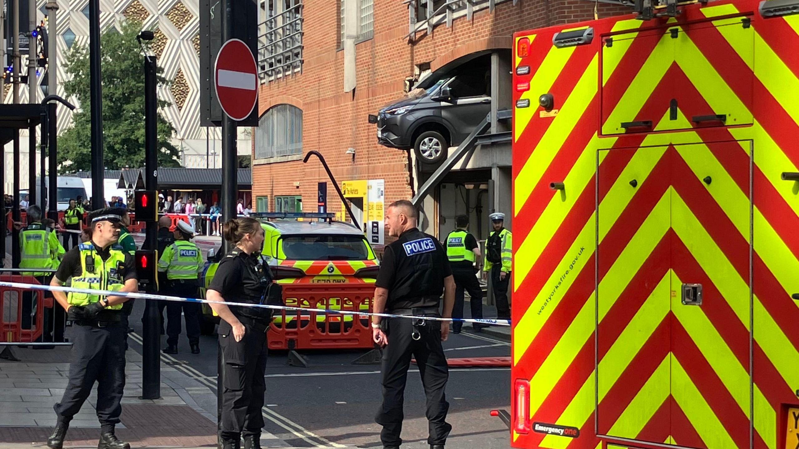 Police officers stand guard next to a cordon, with a car hanging out of the first storey of a large car park in Leeds city centre