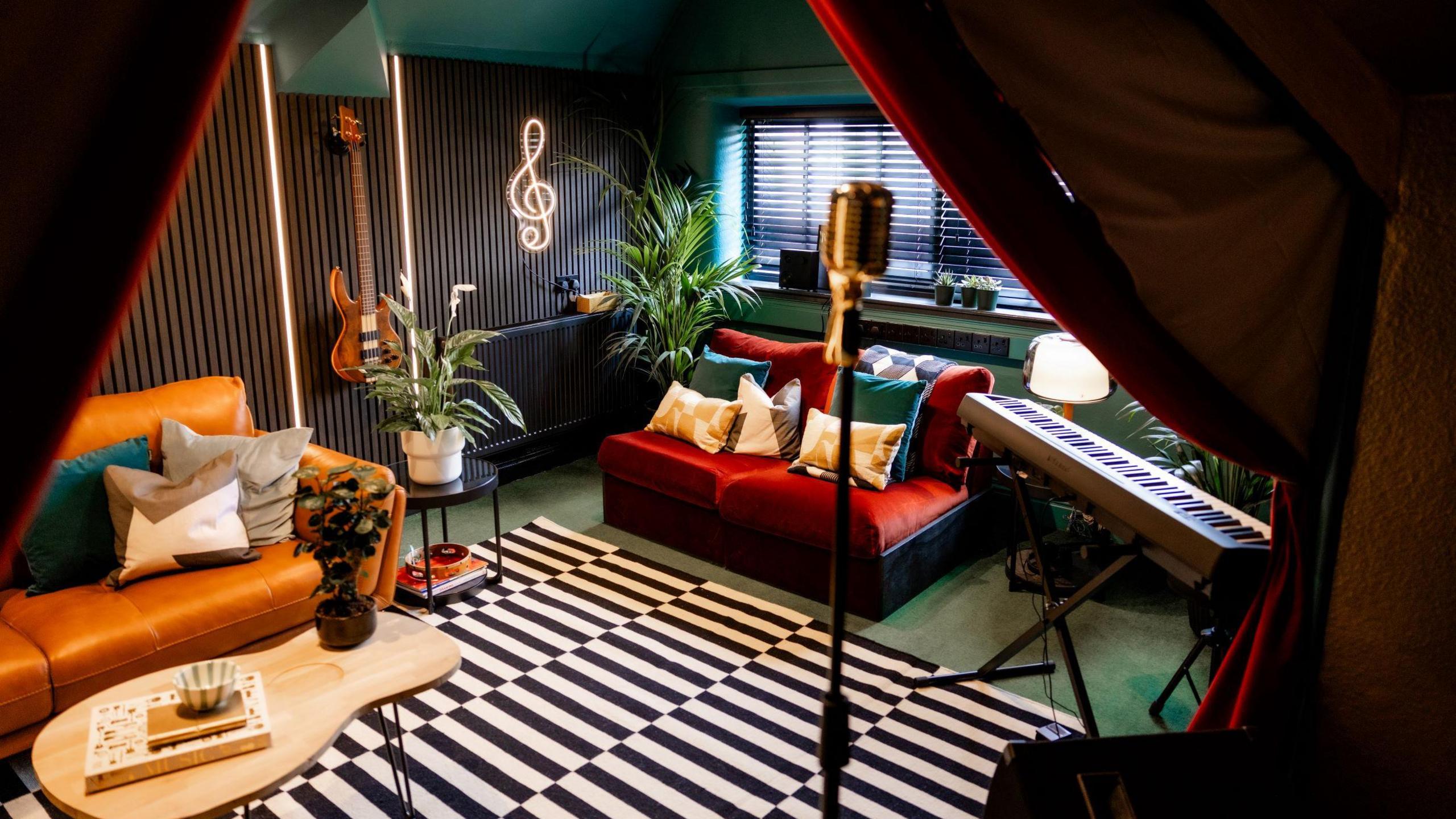 A music room with guitars hung on the walls, which feature wooden slats. On the floor there is a black and white rug. There are two small sofas towards the back of the room, and a keyboard and microphone in the foreground