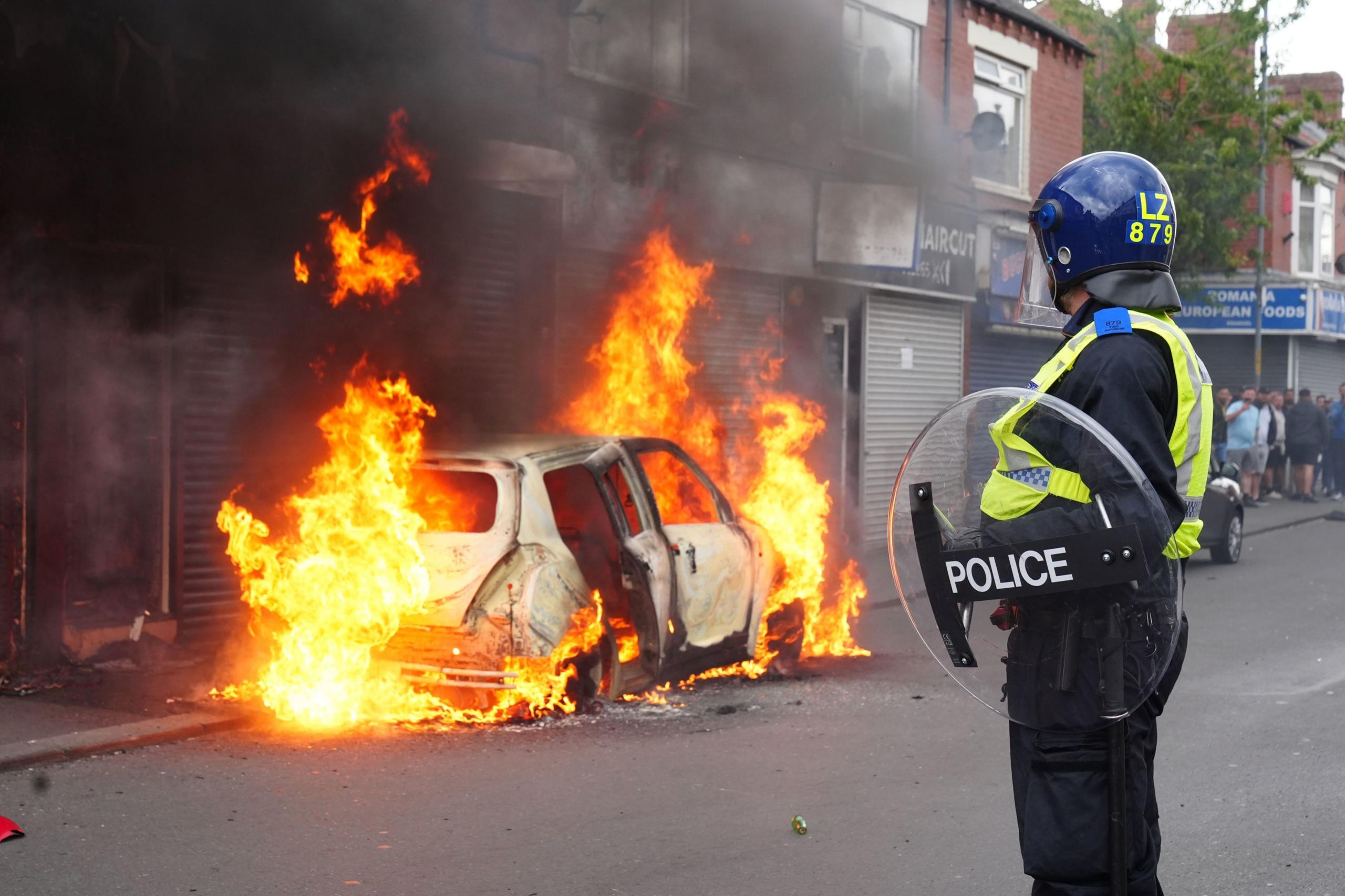 A police officer carrying a riot shield looks on as a car is ablaze on the opposite side of a Middlesbrough street