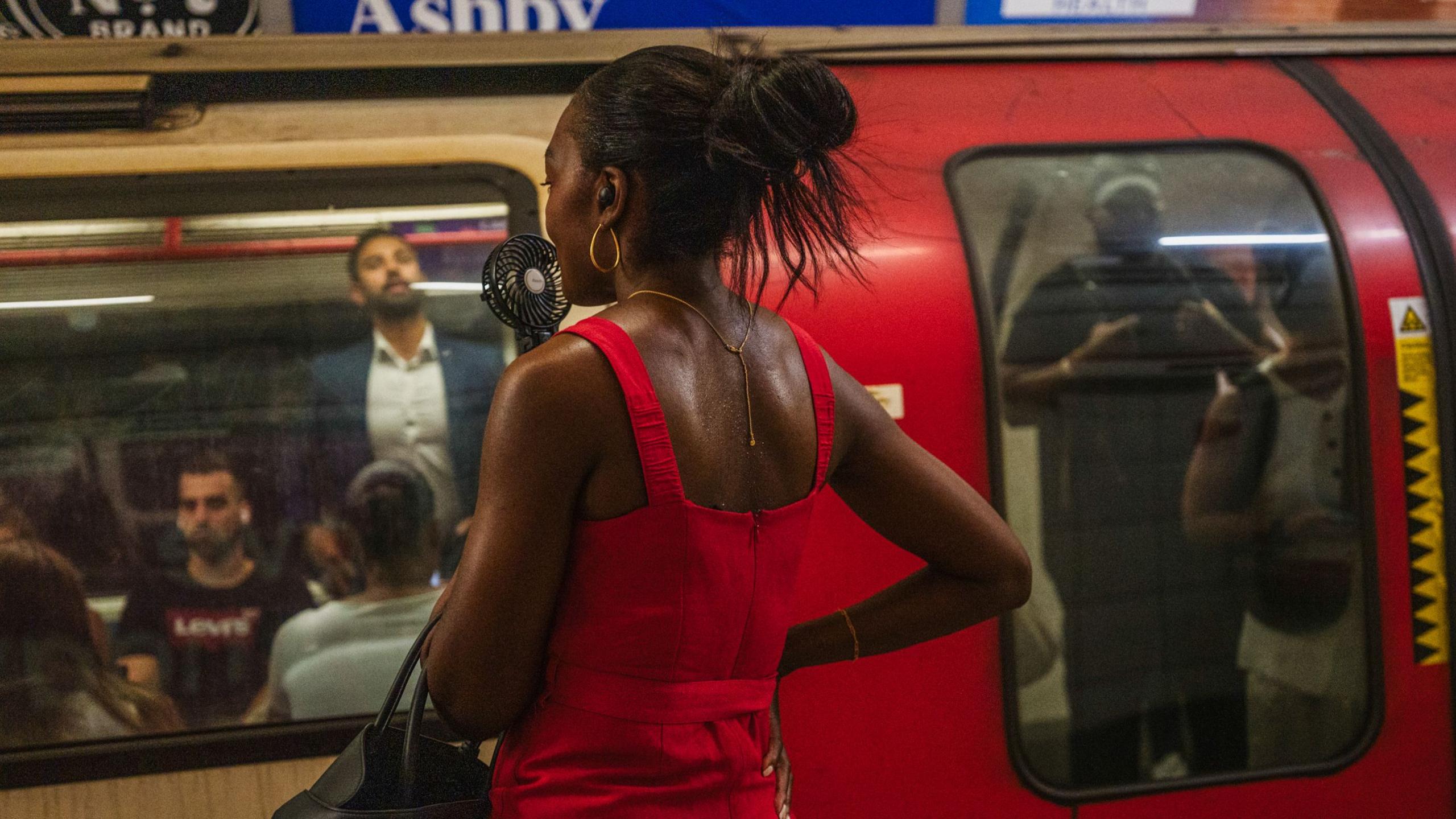 A woman holds an electric fan to her face. She wears a red dress and a tube full of people is in front of her.