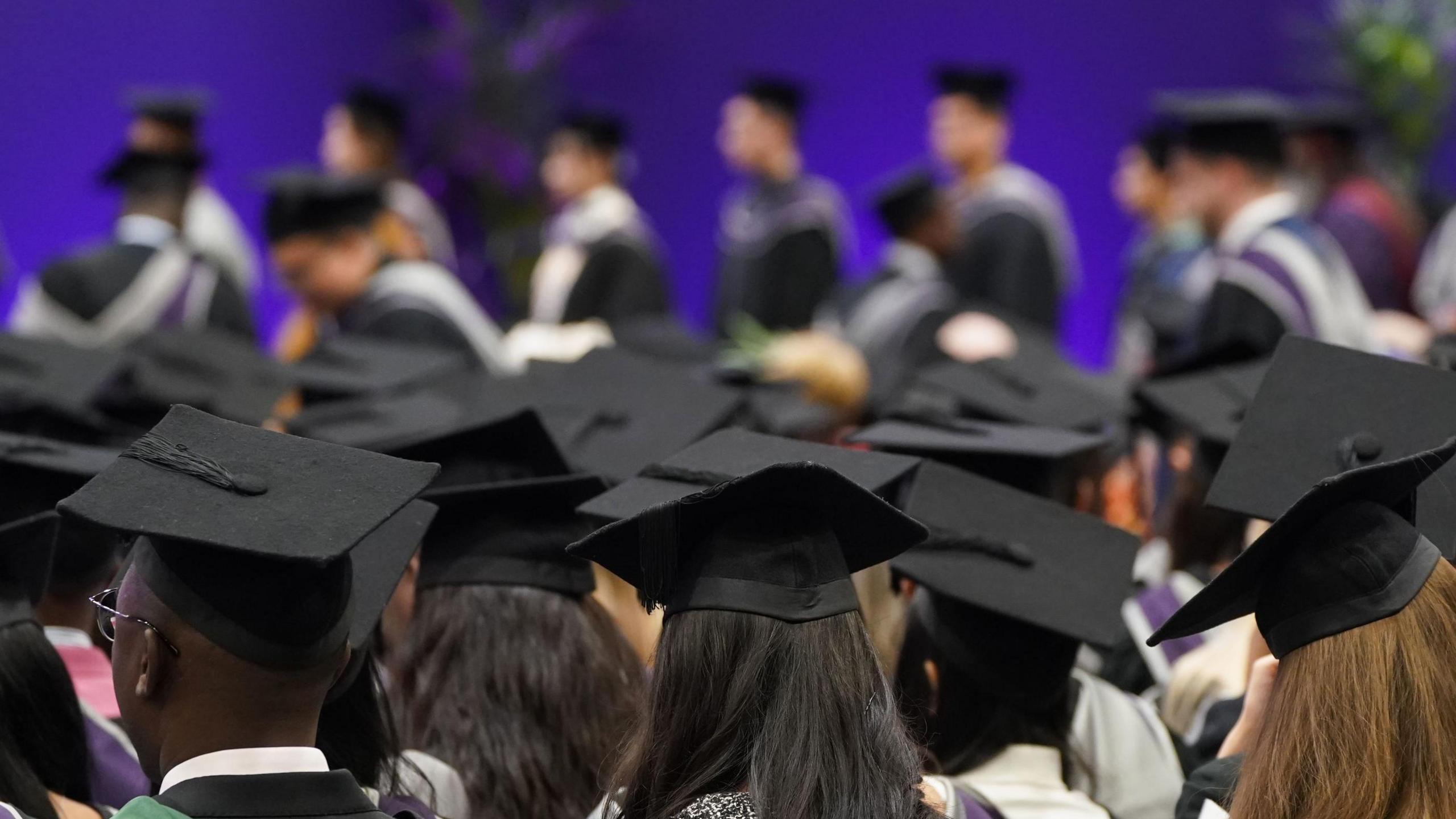 Rows of Degree students wearing gowns and black mortarboards. The background in the distance is purple.