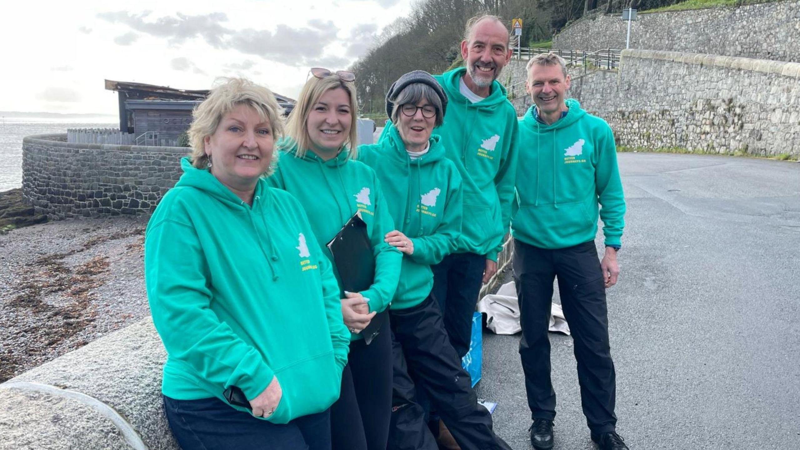 Five members of the group standing in a line. They are wearing matching green jumpers with the group's logo on them. A white image of Guernsey with yellow writing.,