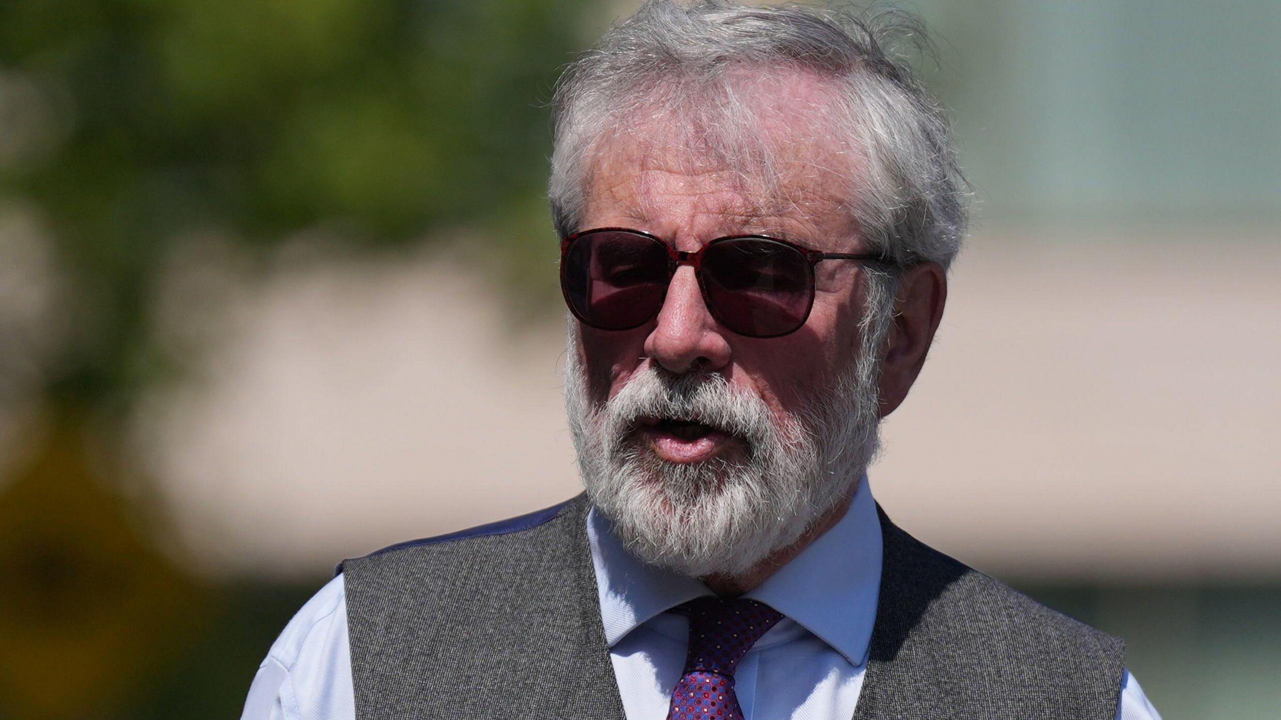 Gerry Adams is pictured from the chest up - he wears a blue shirt, grey waistcoat, dark sunglasses and purple tie