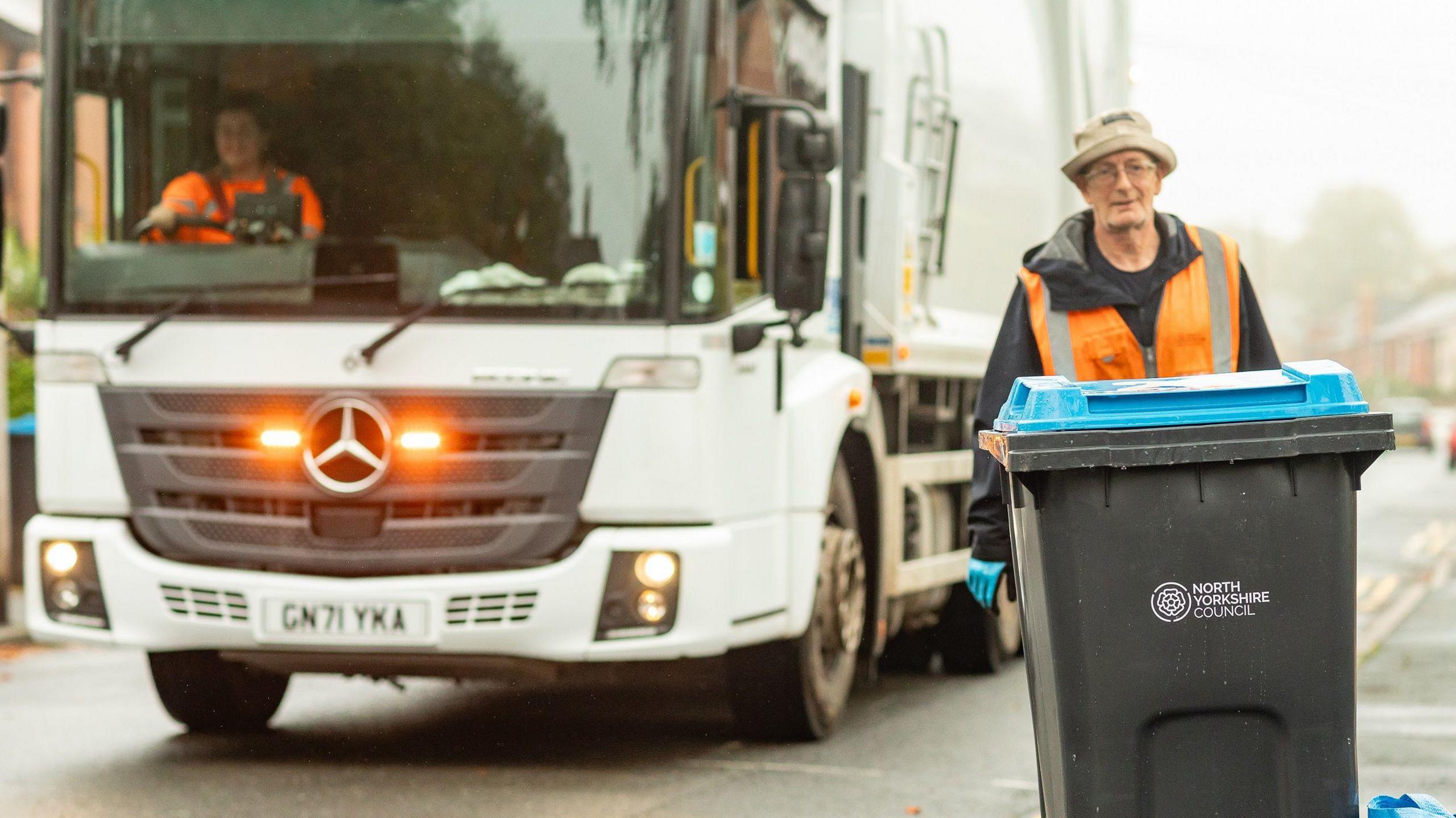 Man with an orange hi-vis jacket wheeling a bin in front of a lorry