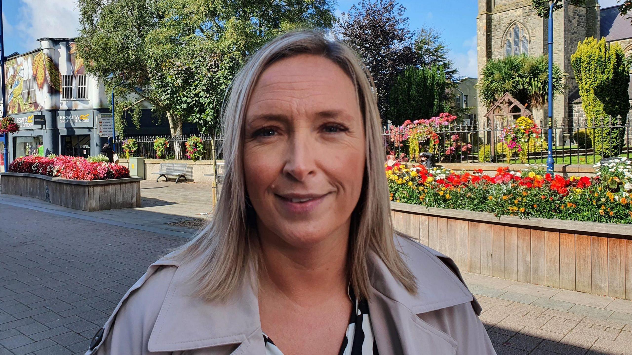 Mandy Corr, in a grey jacket, and with blonde hair, stands in front of a flower bed. She is partly smiling