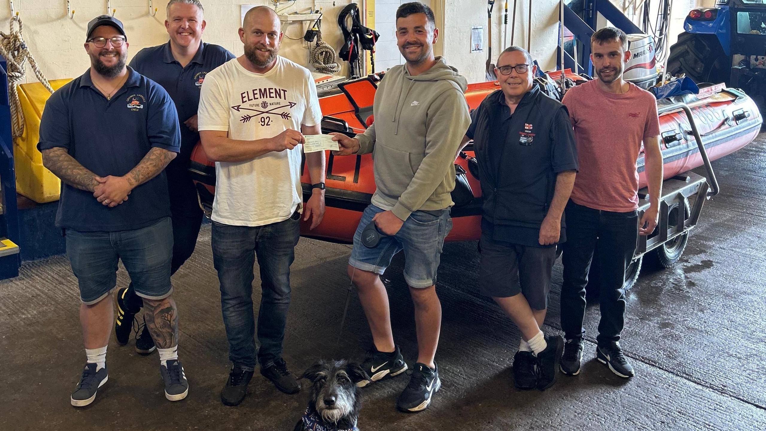 Curtis Johnson is wearing a grey hooded top, denim jean shorts and black trainers, and holding the lead to Rex, a black terrier-type dog. He is handing a cheque to one of the five male members of crew, who are all casually dressed. Three of them are wearing branded RNLI polo shirts. They are standing in the lifeboat station in front of the inshore lifeboat and everyone is smiling.