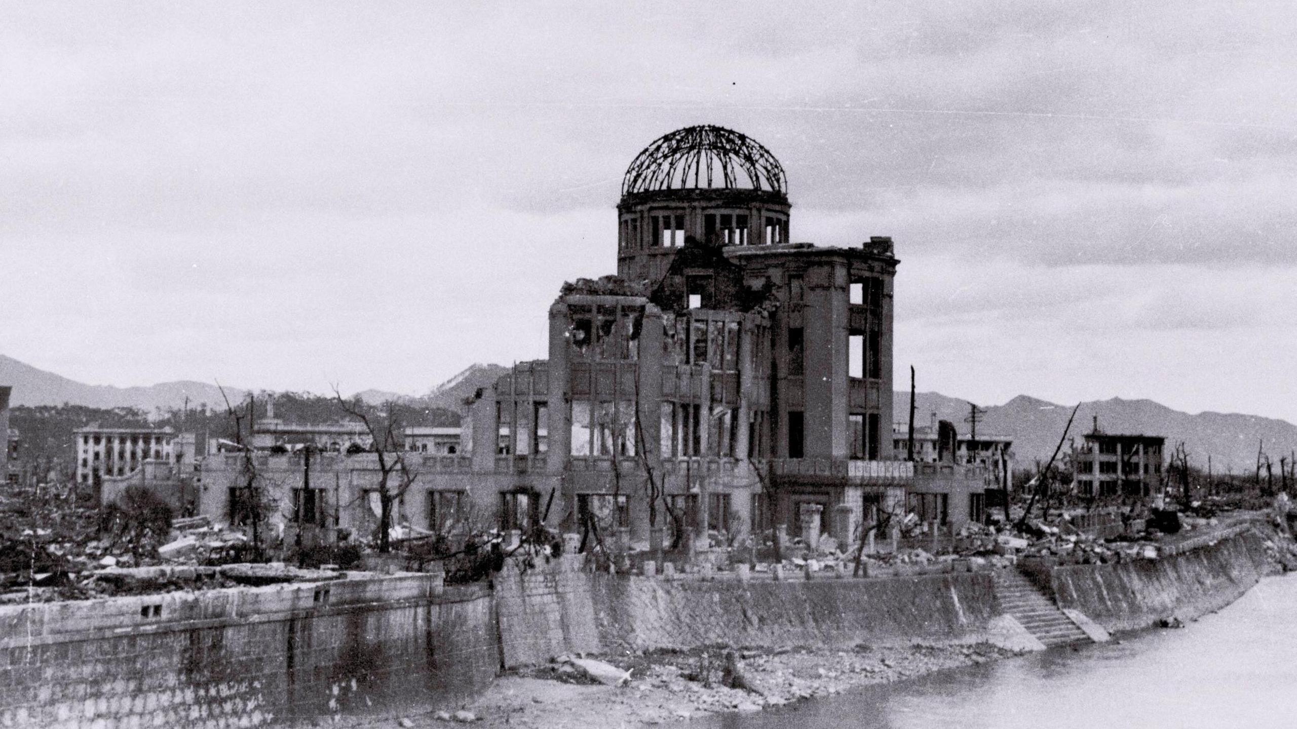 A black and white photo of the damaged remains of the Hiroshima Prefectural Industrial Promotion Hall, where a large domed roof with just the beams remaining sits on crumbed walls. The building sits on the side of the river, with other ruins of building behind it. 