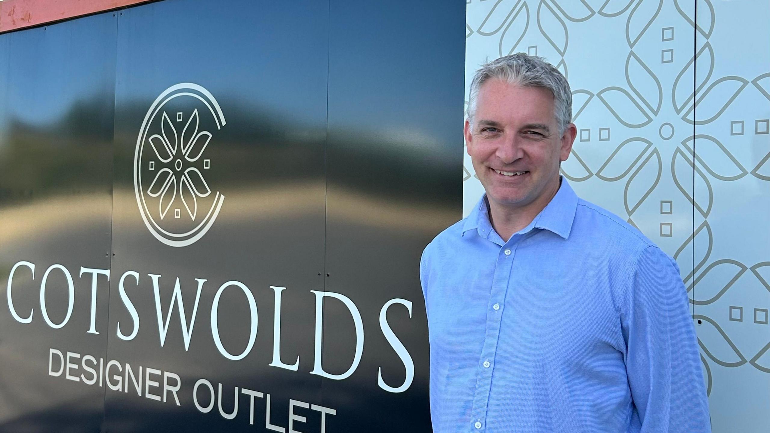 Adrian Field is standing in front of a sign for the Cotswolds Designer Outlet. He is wearing a blue shirt and is smiling.