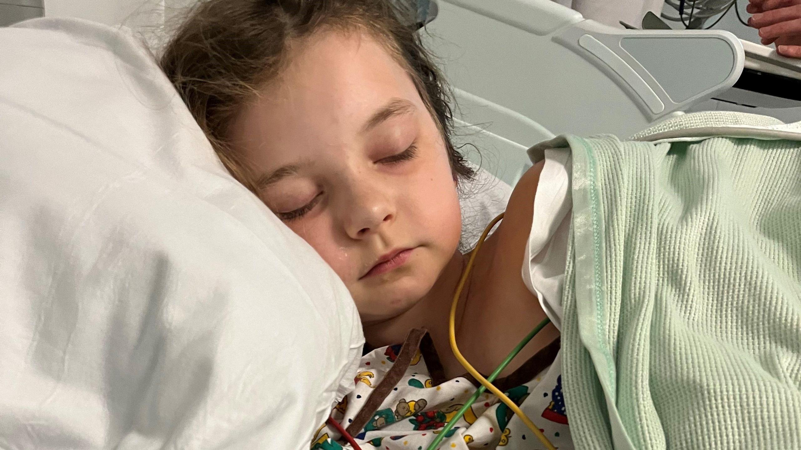 Ivy lying in a hospital bed asleep. She is covered with a light-green blanket and there are some tubes around her chest area. 