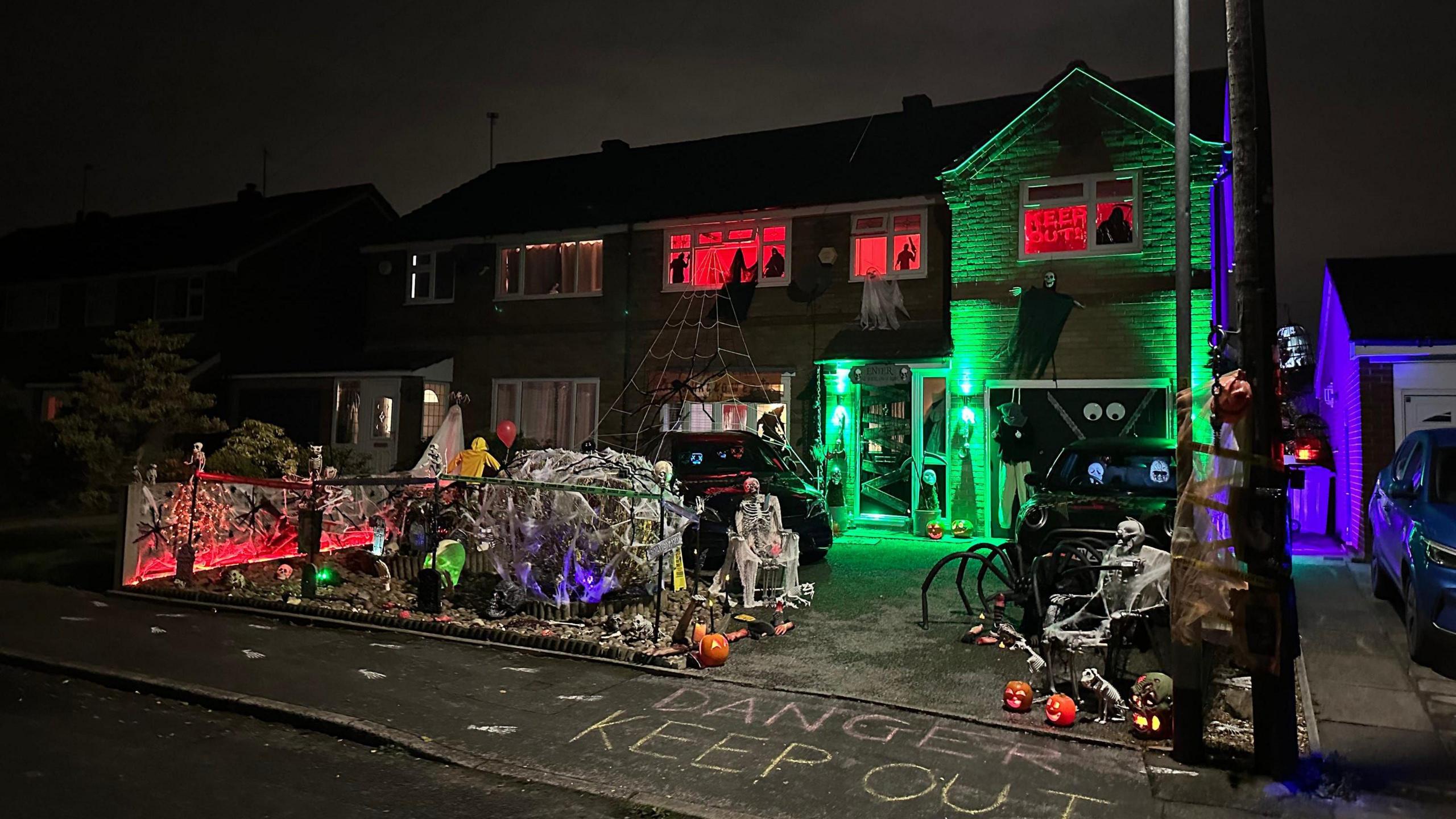 Their home in the dark. The path in front of the house says 'danger keep out' and their front garden is full of lit pumpkins, fake skeletons, cobwebs. The front door is lit green and the windows upstairs are lit red. 