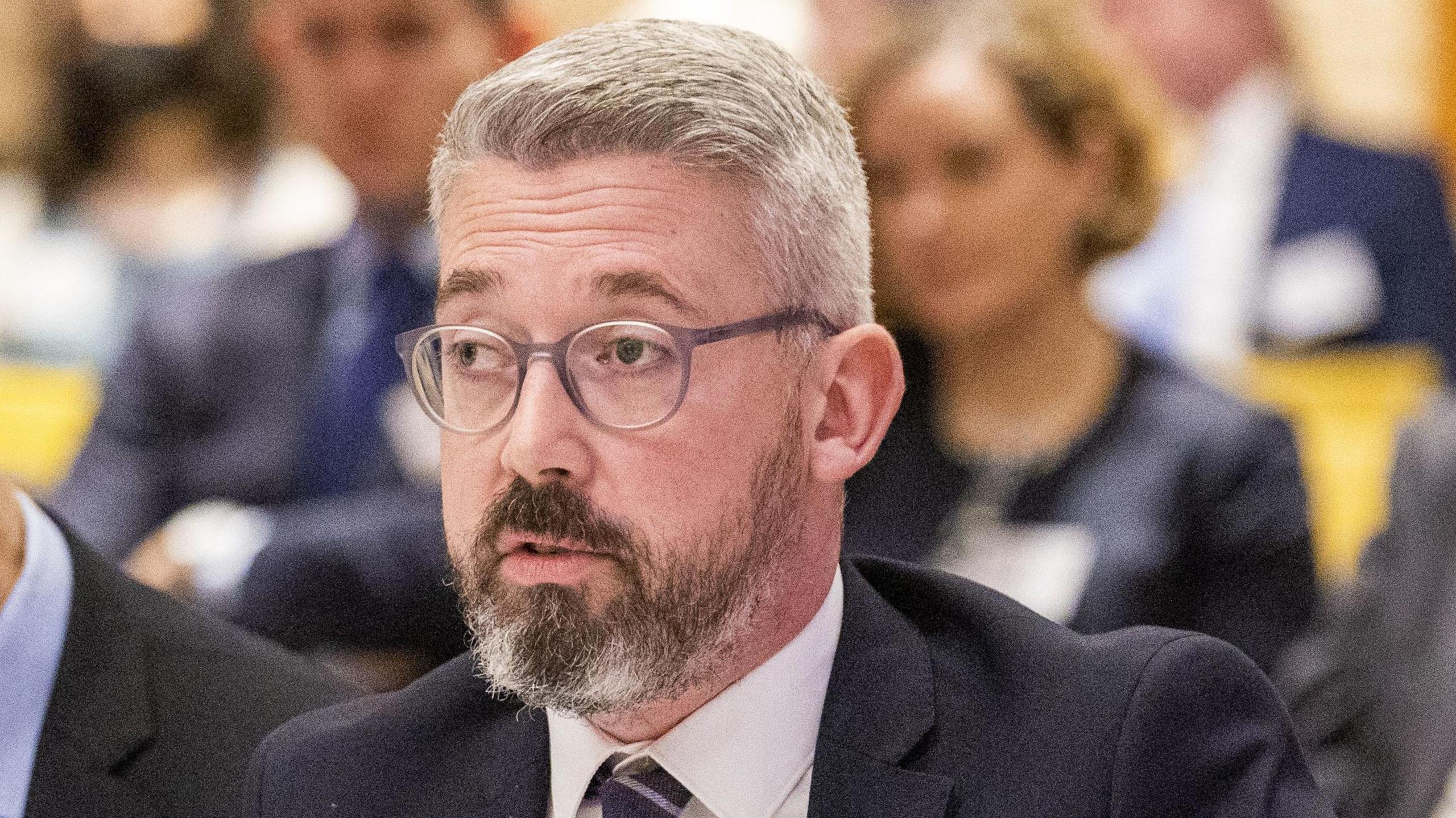 Niall Ó Donnghaile pictured in October 2022. He is wearing thick-rimmed glasses, and he has a greying beard and grey hair.   He is wearing a dark suit, a white shirt and a dark striped tie