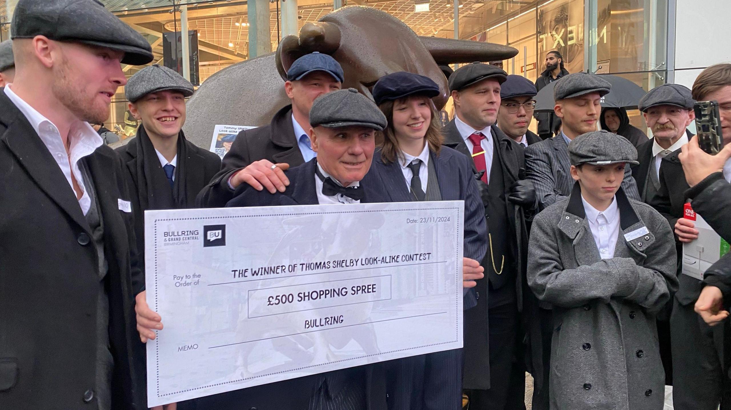 men and women dressed in flat caps and overcoats in front of a bronze bull statue. A man is holding a big white cheque that says £500 shopping spree.