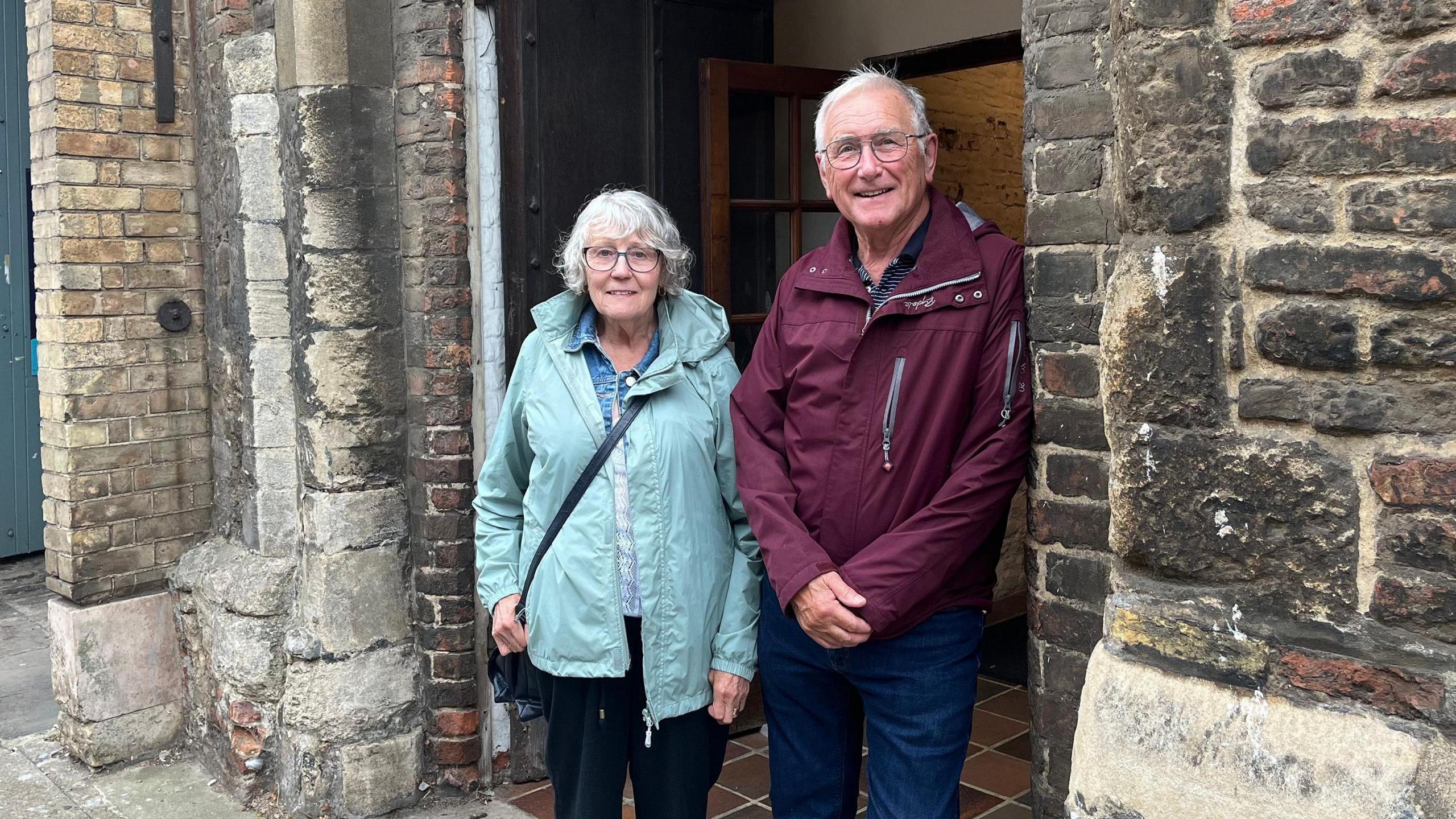 Susan and Roger Brown are standing in a doorway of the medieval Guildhall doorway. She is wearing a teal raincoat and black trousers and has grey bobbed hair. He is wearing a crimson raincoat and blue jeans. Both wear glasses.