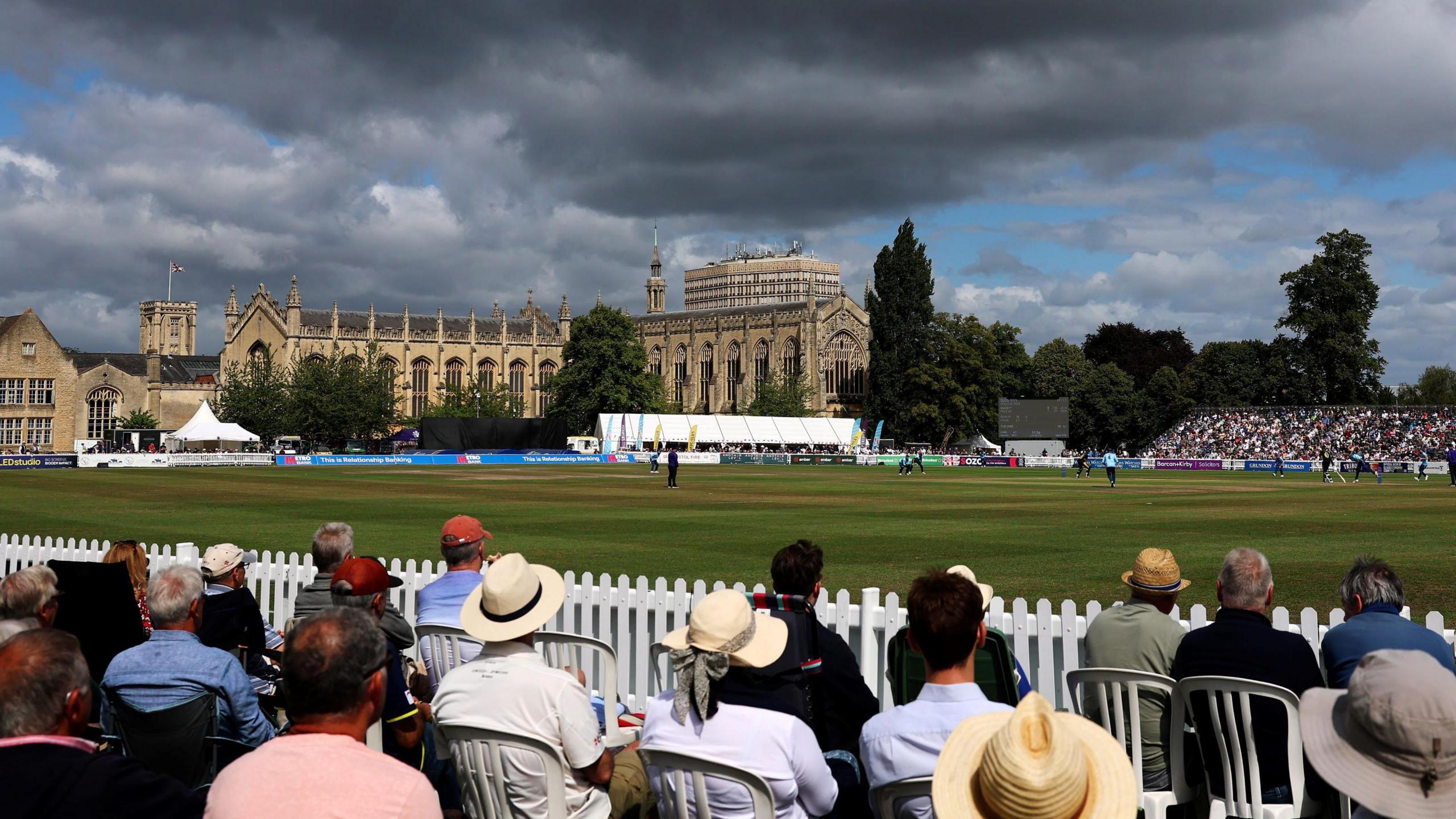 Cricket fans sit on seats on the boundary edge at the College Ground in Cheltenham as Gloucestershire play a home game. Between the spectators and the playing surface is a white fence, about waist-high. Another grandstand is visible in the distance, and it is full of spectators. The grand college buildings are also visible, and there are dark grey clouds against a blue sky.