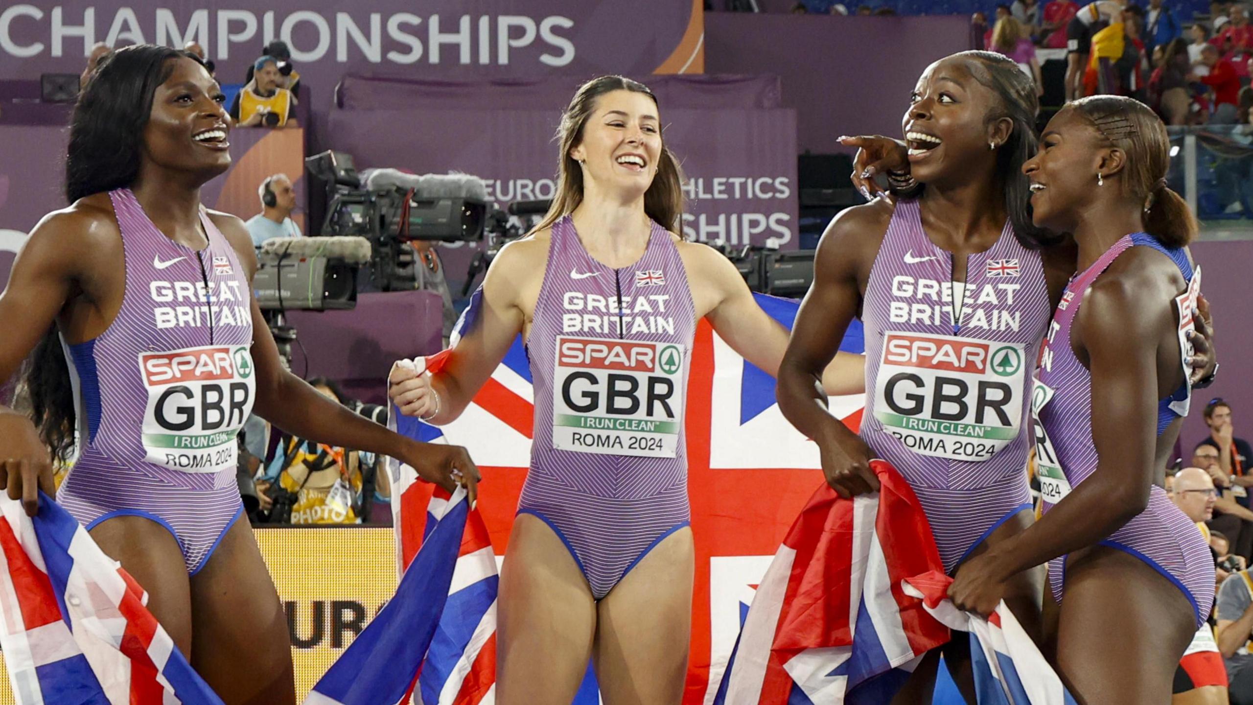 Four female athletes (Daryll Neita, Amy Hunt, Desiree Henry and Dina Asher-Smith) wearing purple Great Britain vests look happy and drape themselves with Union flags after winning gold medals in a relay race