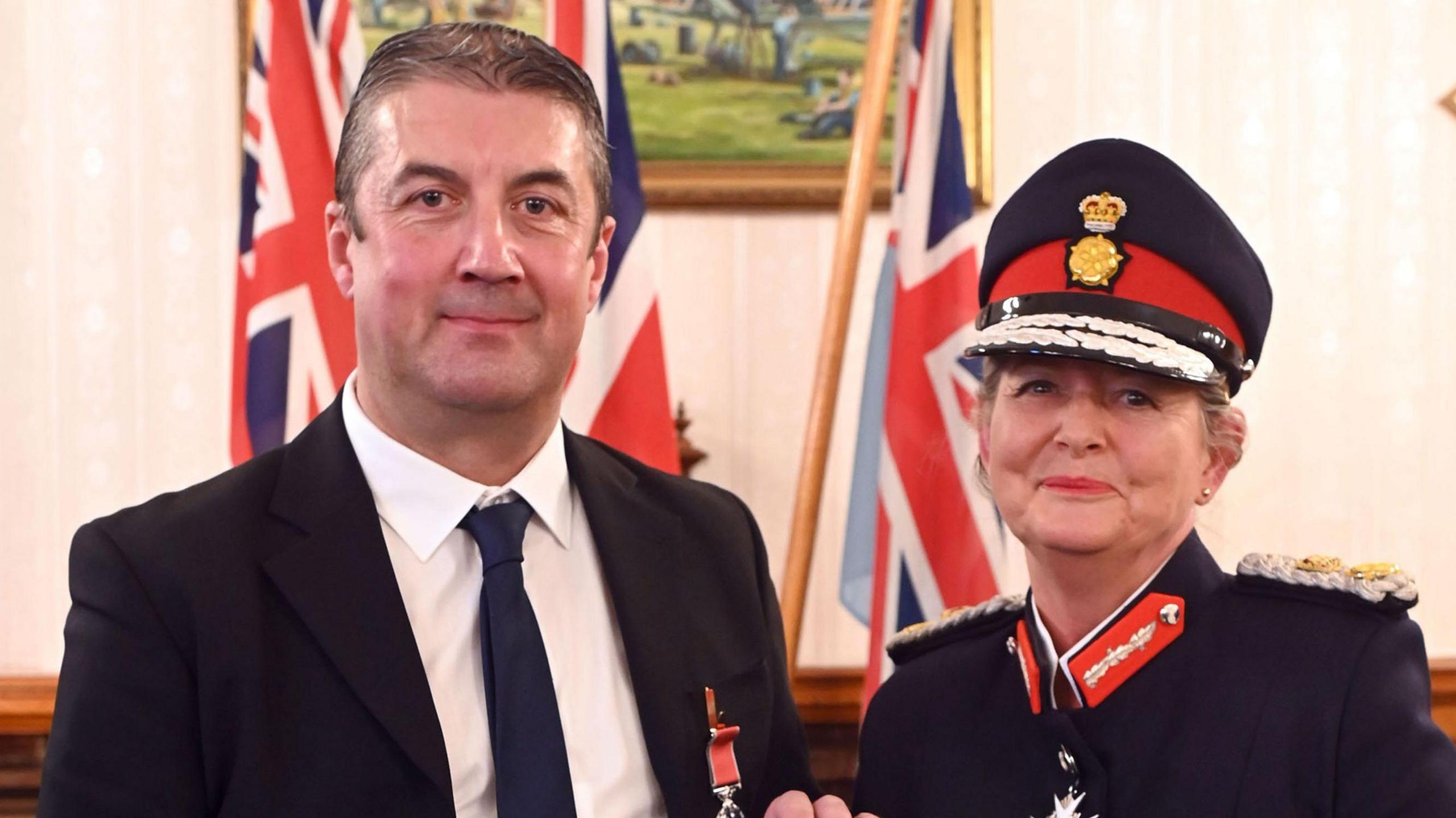 Joe Healy, in a dark blue suit with slick back hair, standing next to Susan Lousada, in full regalia with red lipstick on. In the background there are three union flags, with a painted picture of a meadow on the wall in the middle. 