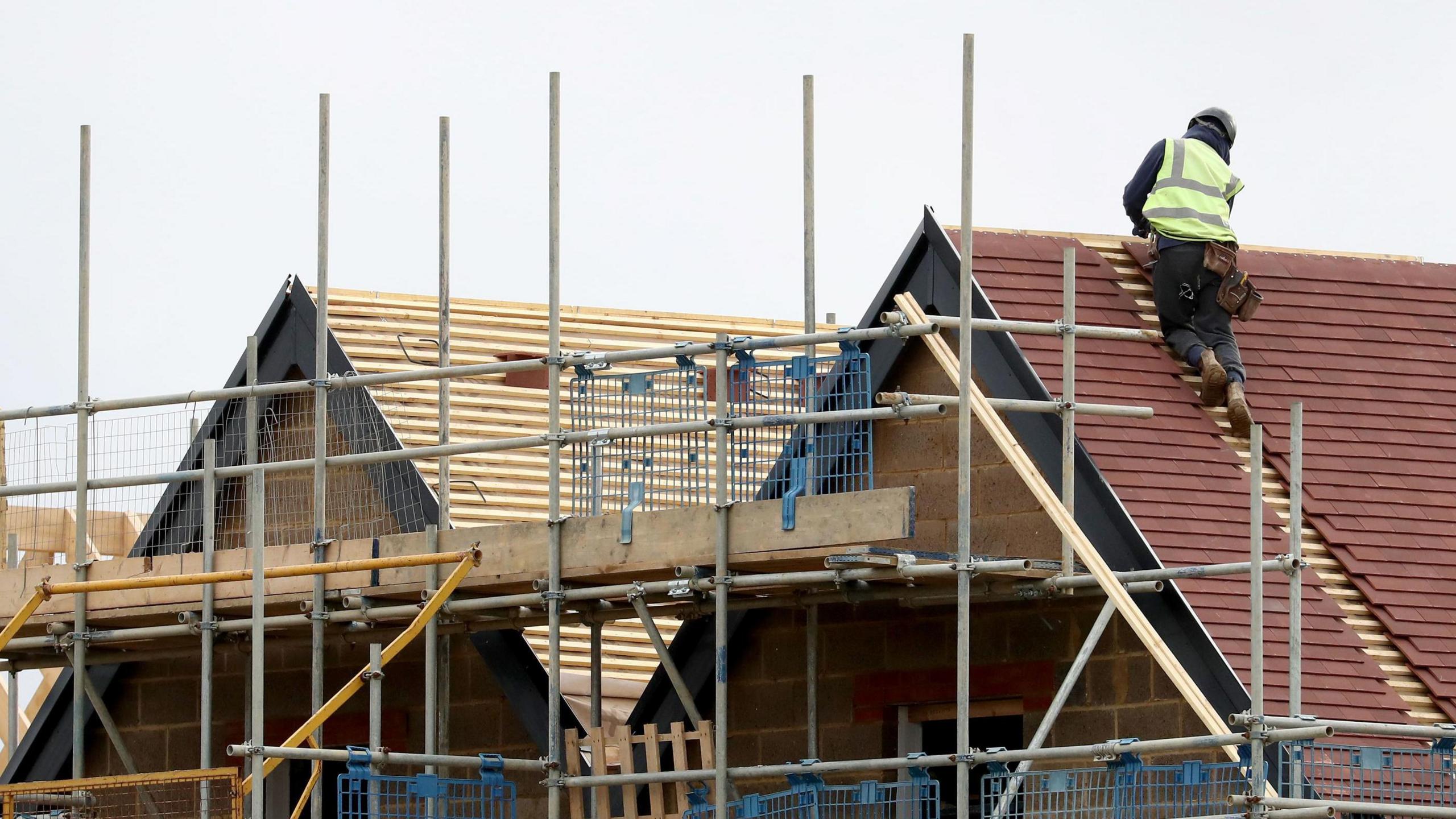 Houses under construction with scaffolding surrounding them.  A workman in high vis clothing is on one of the roofs.