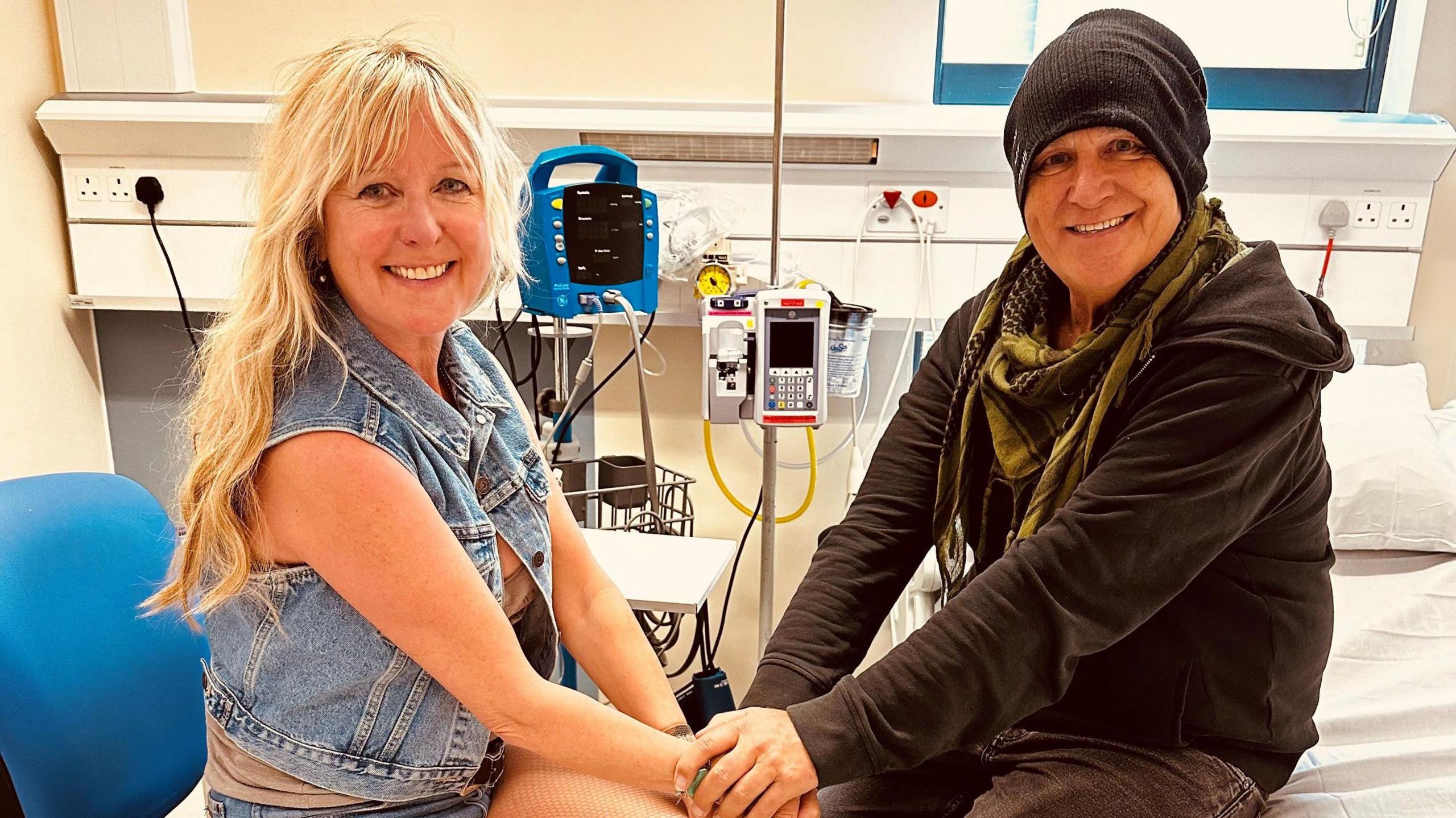 Mike Peters sat in a hospital bed. He wears a khaki scarf and black zip hoodie, with a black beanie hat on his head. His wife sits on a blue chair on the side of the bed and the pair hold hands in front of their knees. She has long blonde hair and a full fringe and wears a denim waistcoat. The pair smile widely towards the camera. 
