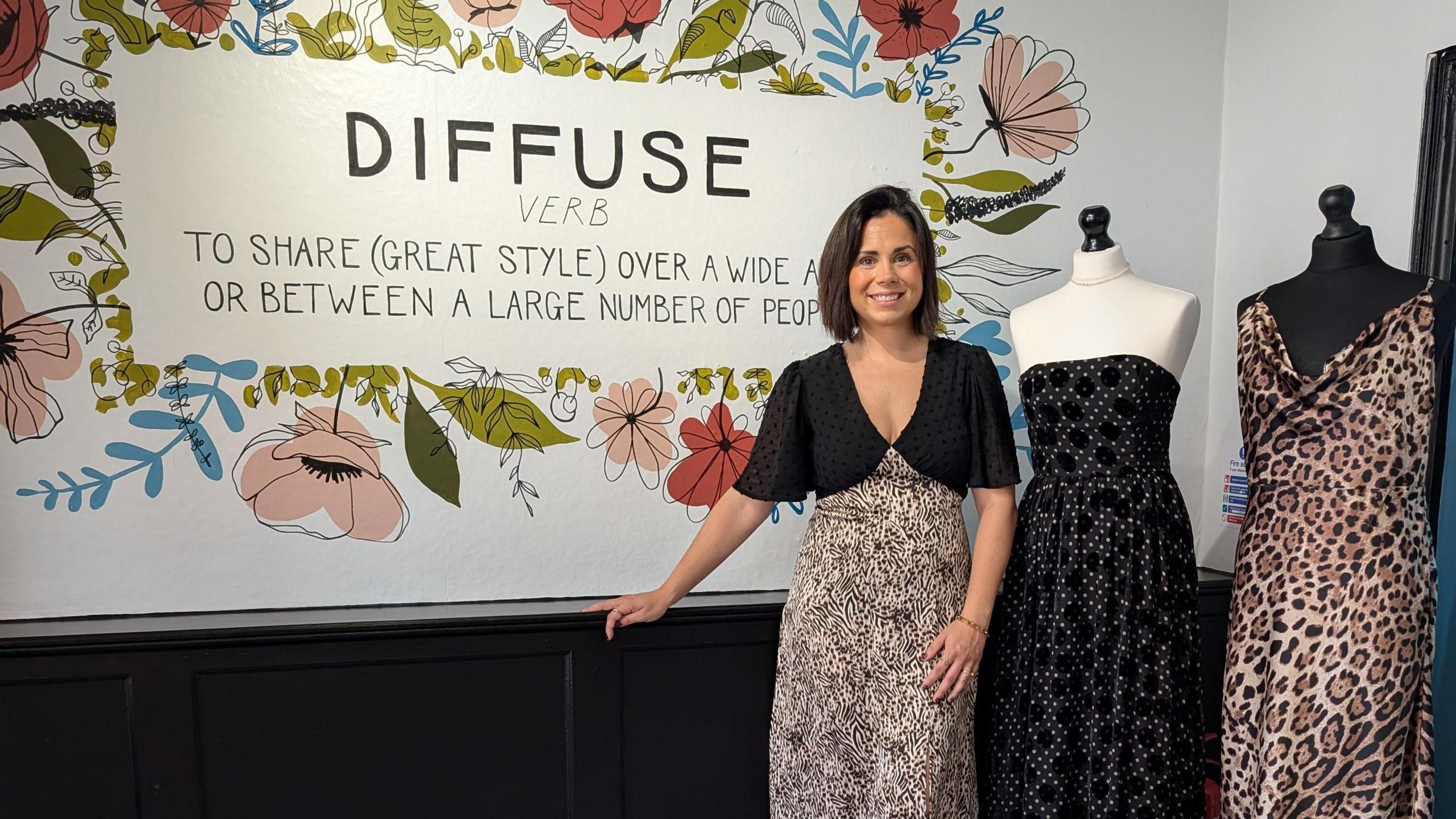 A woman wearing a black top and skirt stands in front of a sign that reads 'Diffuse'. To her right are two mannequins, each with a dress on.