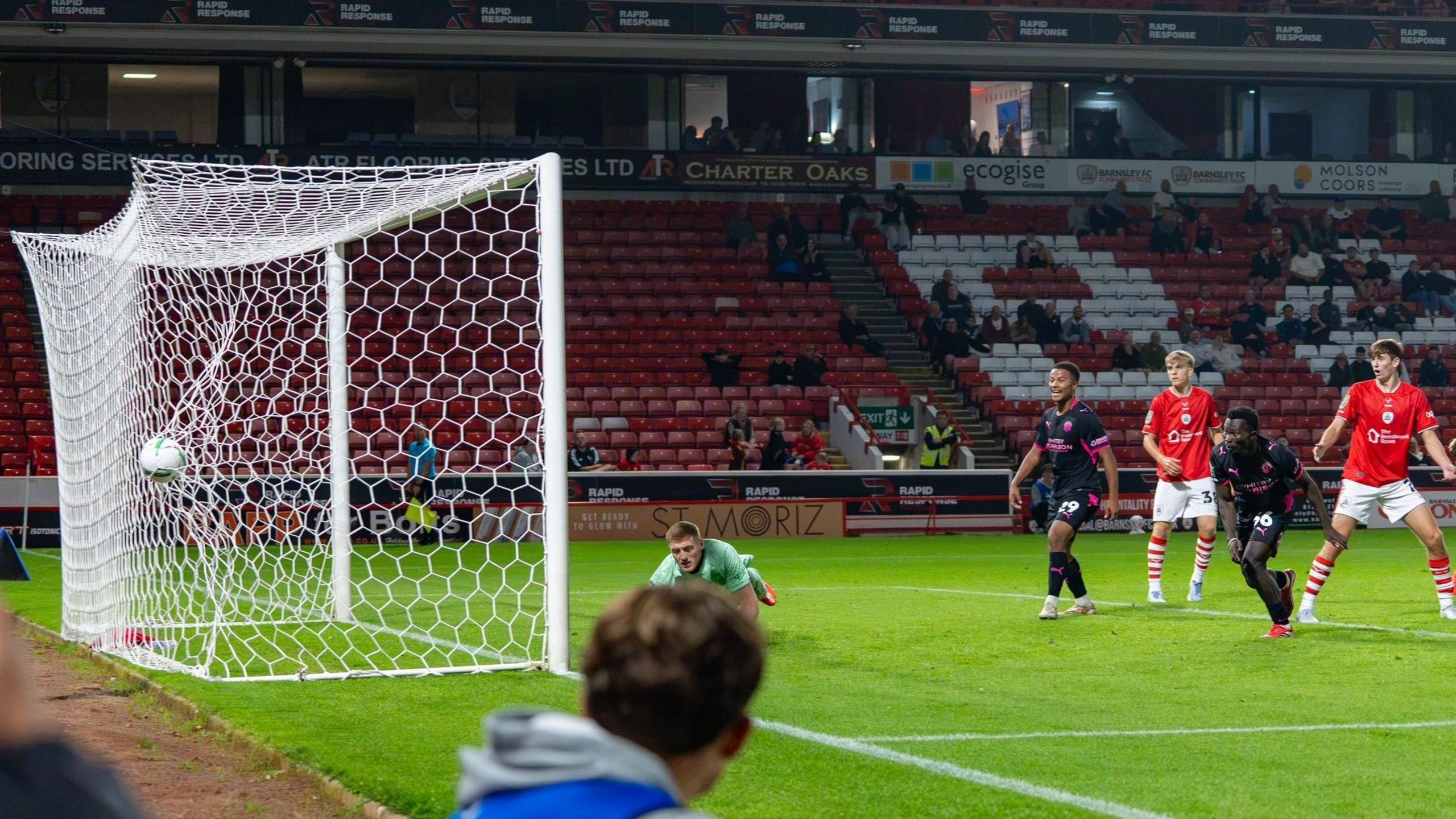 Joel Ewusi (second from the right) watches as his header goes past the goalkeeper and into the net. He is wearing a black kit and is standing between two Barnsley defenders in red shirts and white shorts. There is another Crewe player between the goalkeeper and the first Barnsley player.