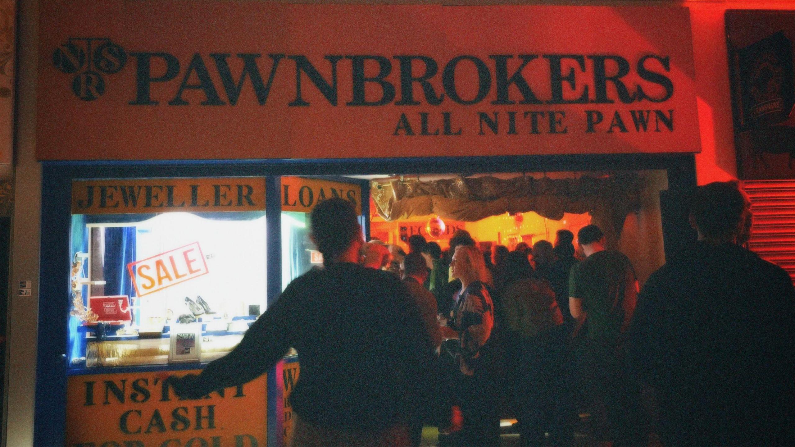 People queue outside a pawnbrokers, it has a yellow and blue sign and adverts for jewellery and instant cash in the window
