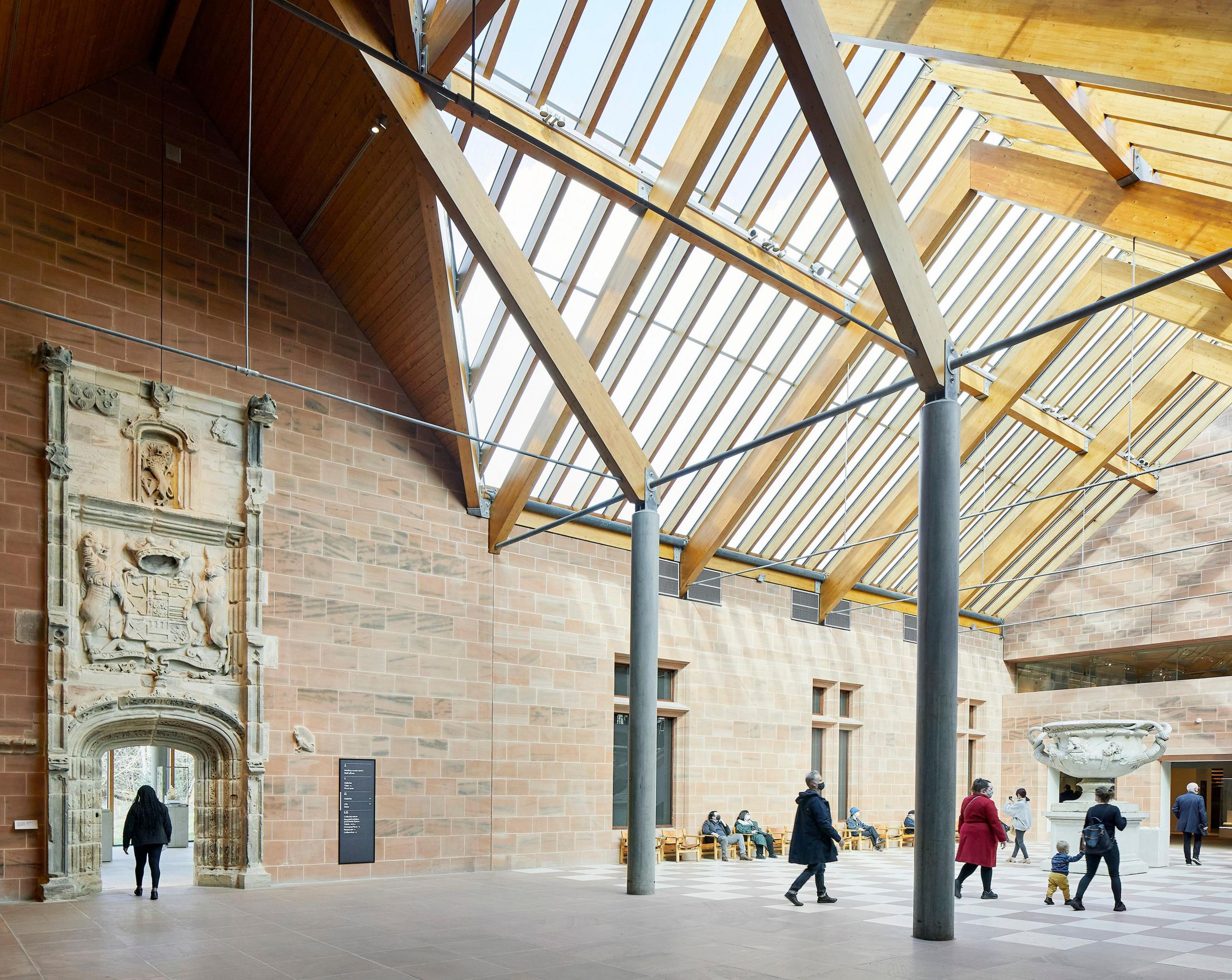 A large open space within the Burrell Collection - with an intricate carved stone archway on the left, a big space with large grey pillars and a glass-paned roof. There are a few people milling around.