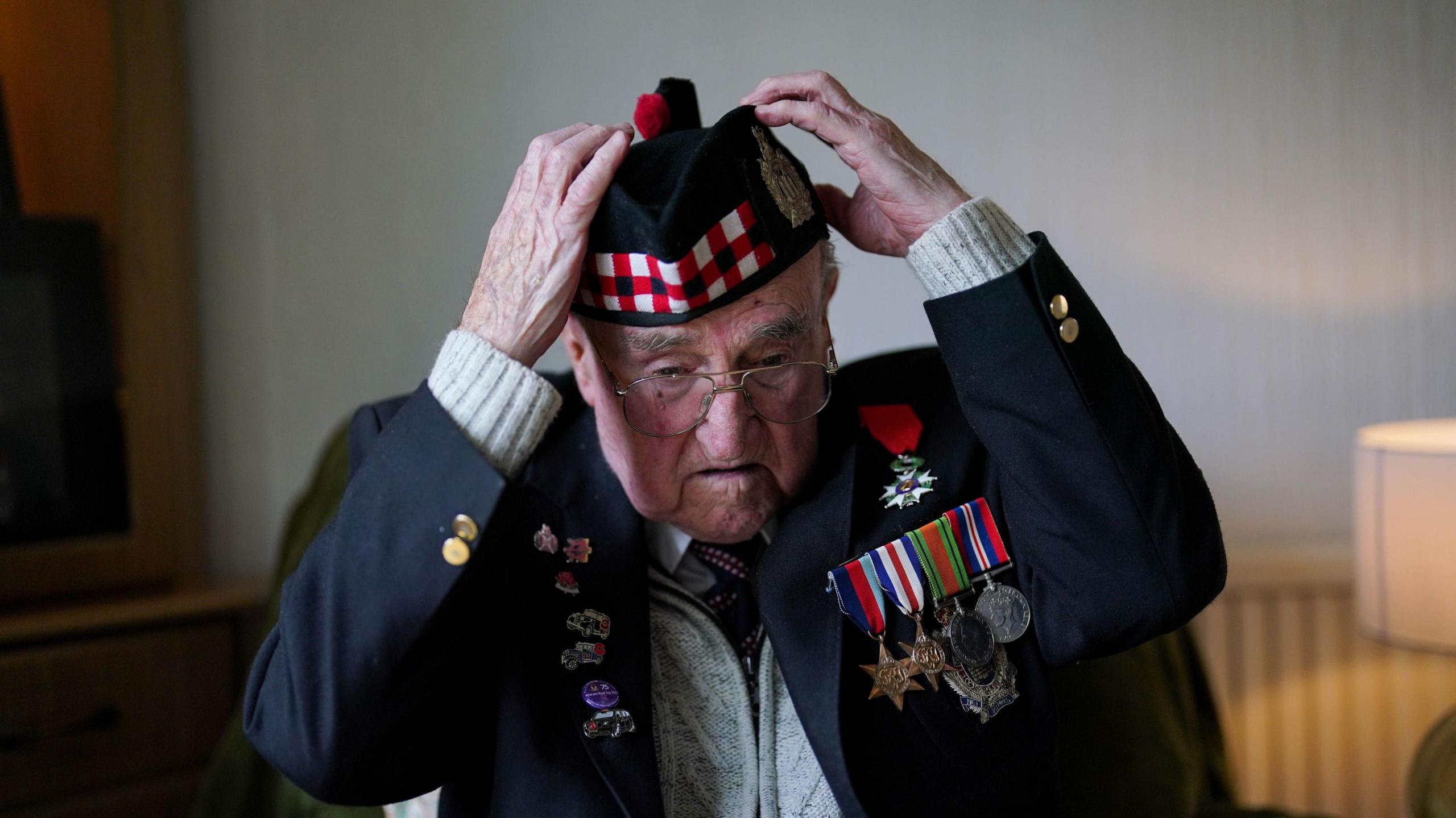 An elderly man is sat in the middle of a room adjusting his hat. He is wearing a navy coloured blazer that has multiple war time medals on it's chest. His hat is also navy coloured but has a red, white and black stripped pattern along the front of it with a red bobble on the top.