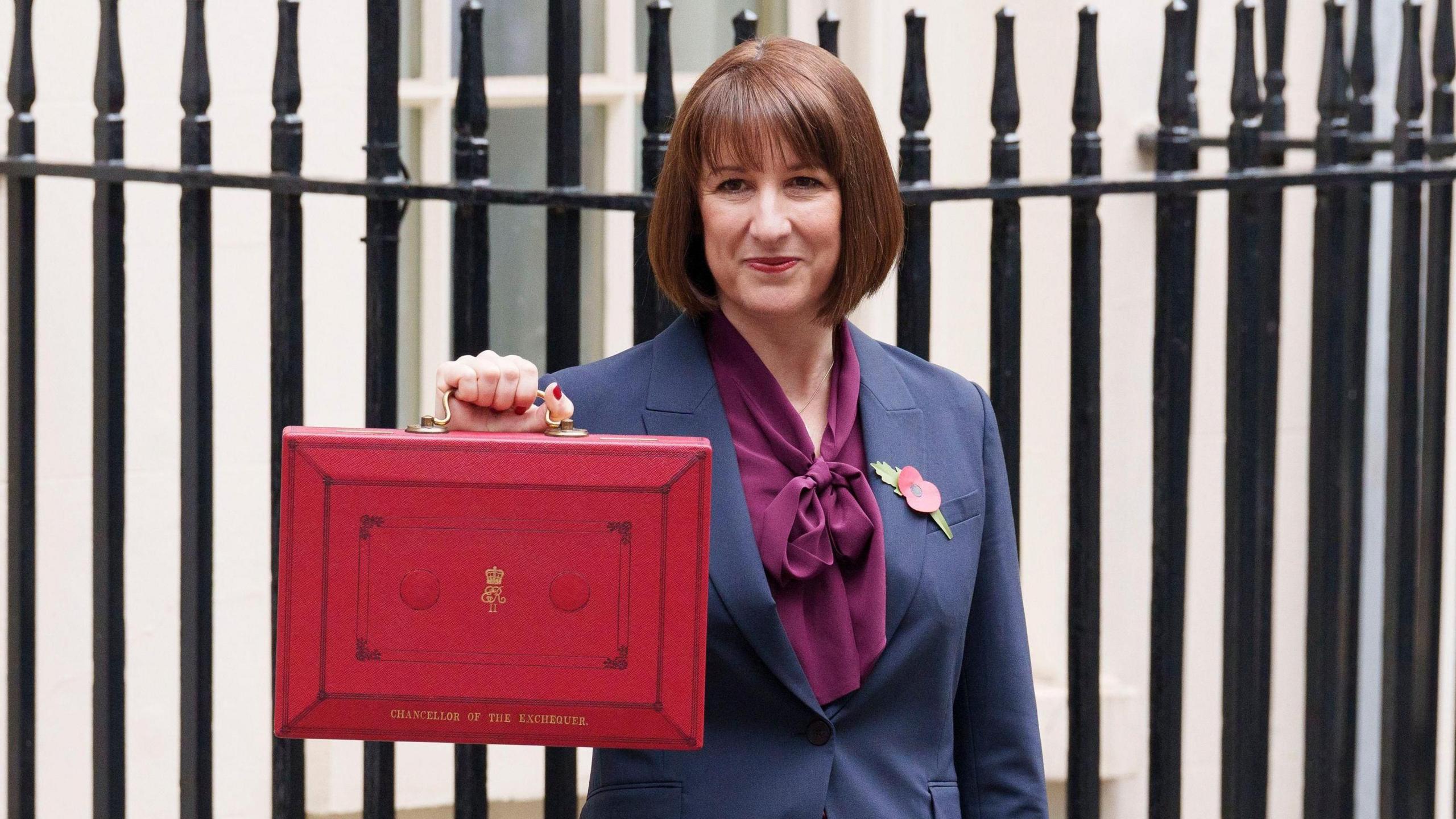 Rachel Reeves holds the red budget briefcase in Downing Street. She has short brown hair and a fringe which frames her face. Ms Reeves is wearing a navy blue blazer, with a paper poppy on the left panel of her jacket. She has a deep mauve purple blouse on underneath, which has a bow tie collar. Her nails are painted red and she is looking slightly off camera, smiling and she presents the briefcase, it is an A4 size, red, with a gold handle and gold detailing on the front, with the words 'Chancellor of the Exchequer' in gold at the bottom. Ms Reeves stands in front of black railing and a white building behind it.