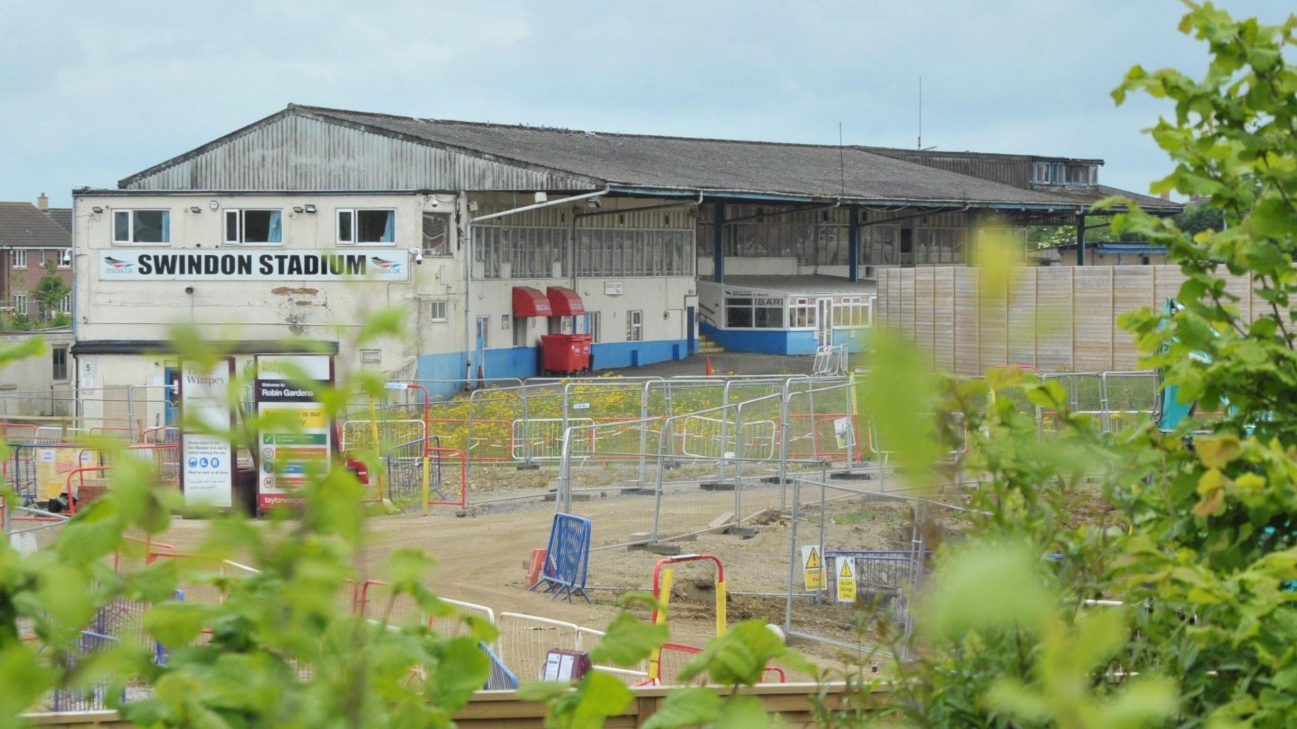 Swindon's Abbey Stadium on Lady Lane. The building's cream paint is coming off and doors are boarded up. It has a corrugated iron roof and the building looks generally run down. There are signs of building work that has been ongoing at the site - with metal fencing around the stadium and a Taylor Wimpey sign near the entrance. The photo is taken through some trees and branches are visible in the foreground.