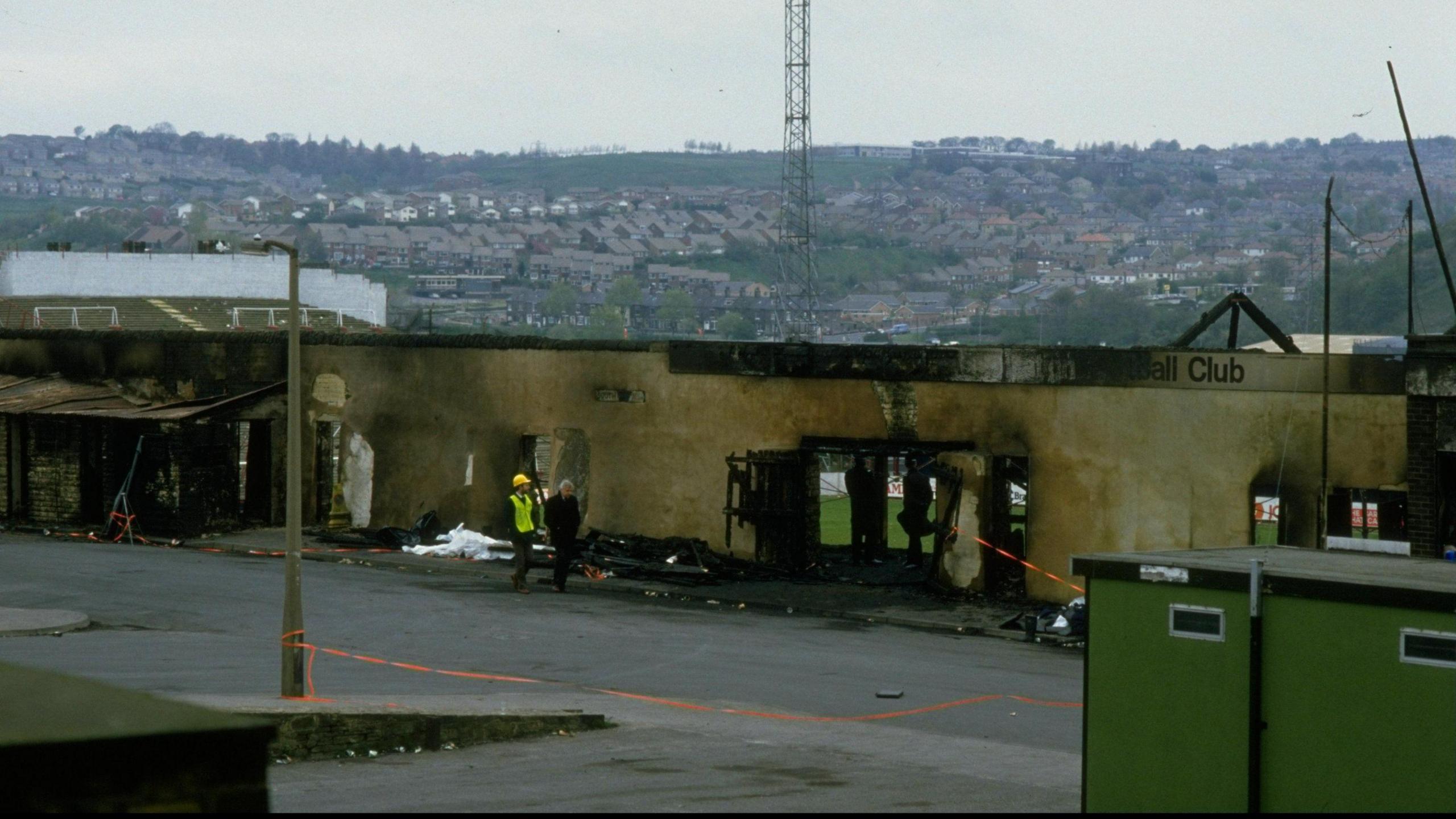 A burnt-out stand at Valley Parade's ground in 1985. Two men, one of whom is wearing a hard hat and a luminous jacket, can be seen walking alongside it.