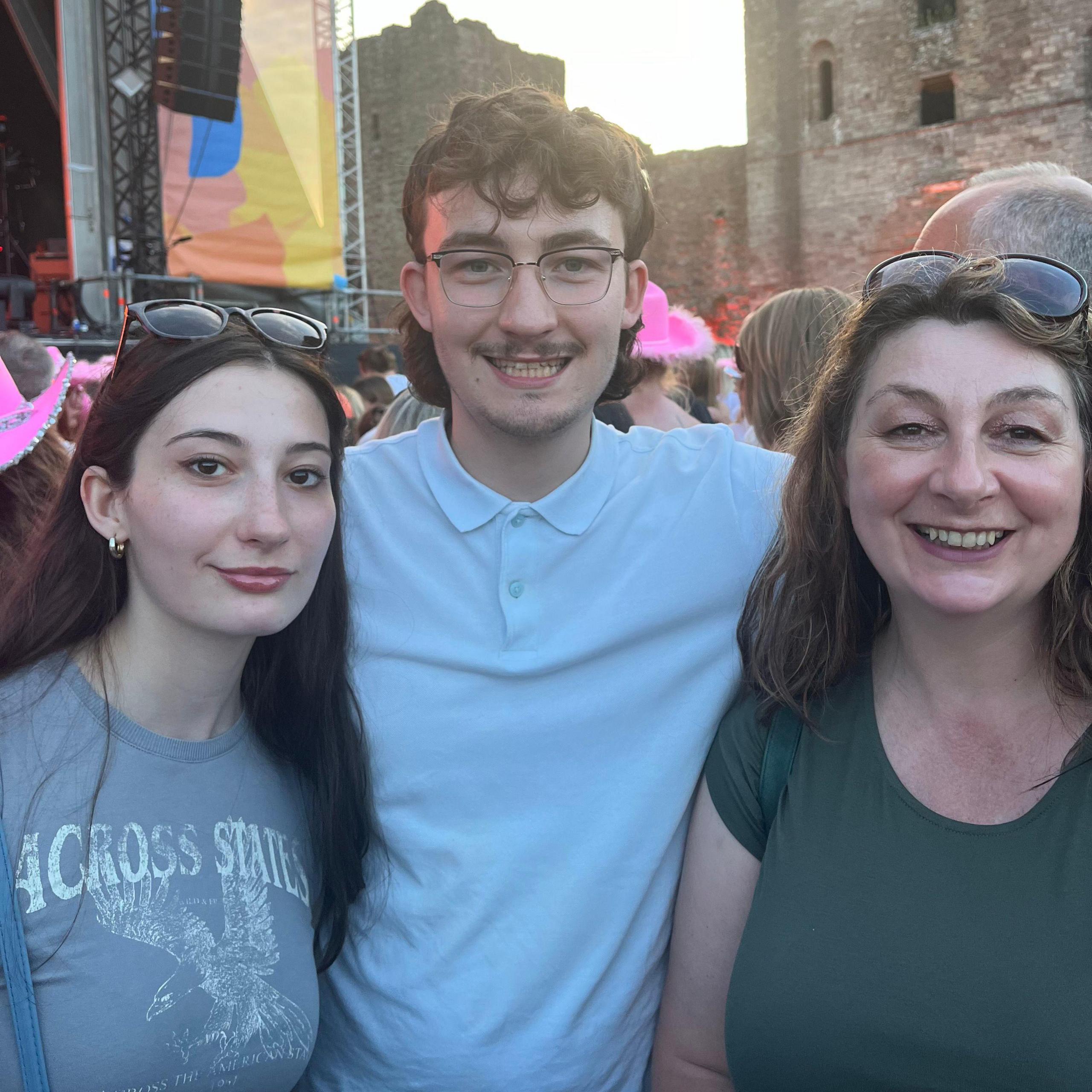 A young woman with brown hair in a grey shirt stands next to a young man with glasses and a light blue shirt, who is also next to a woman with brown hair and a green shirt. The two women, who are a mother and daughter, both have sunglasses pushed up onto their heads