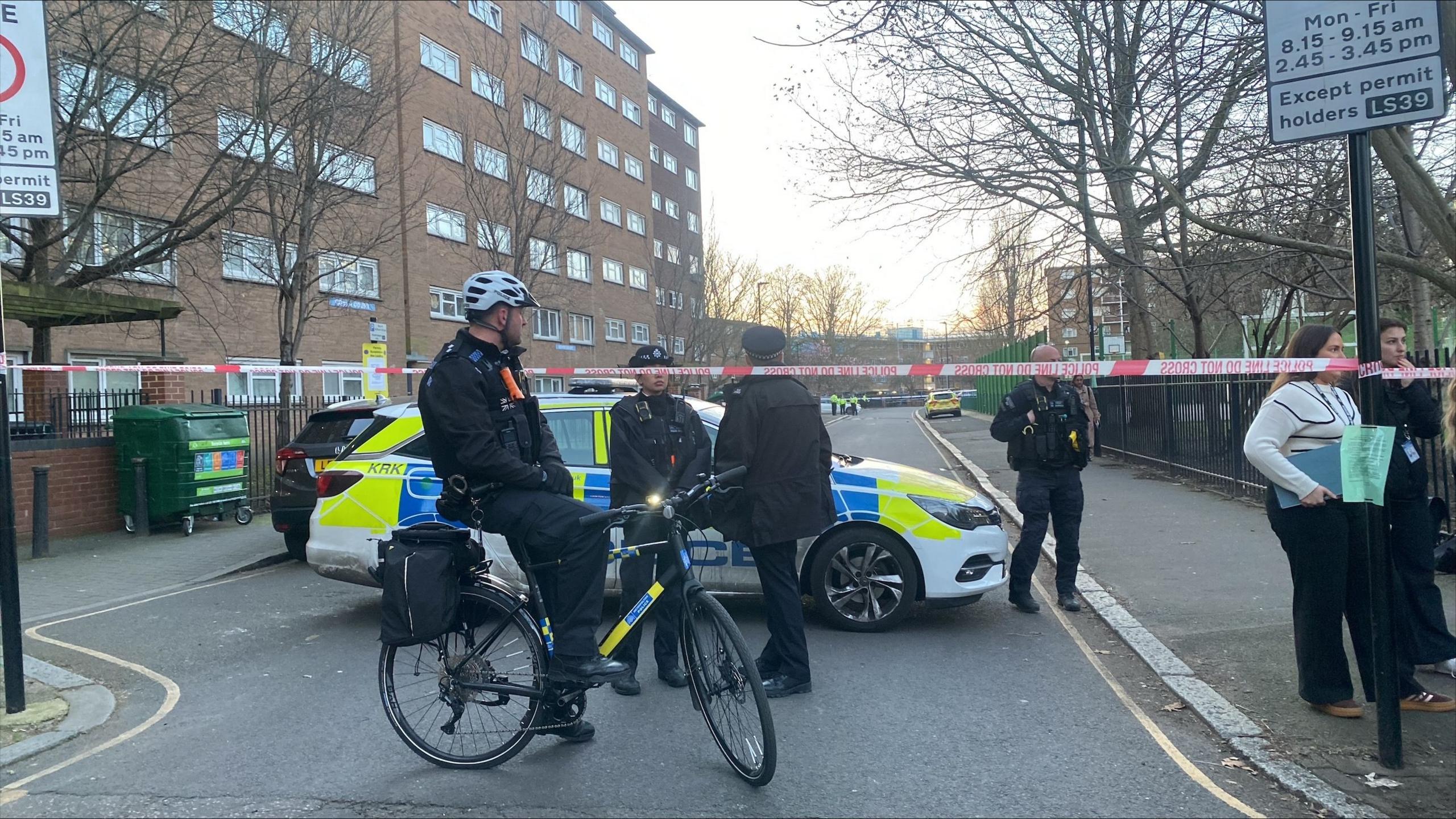 Police car and officer on bike beside red police cordon with further police activity down the road