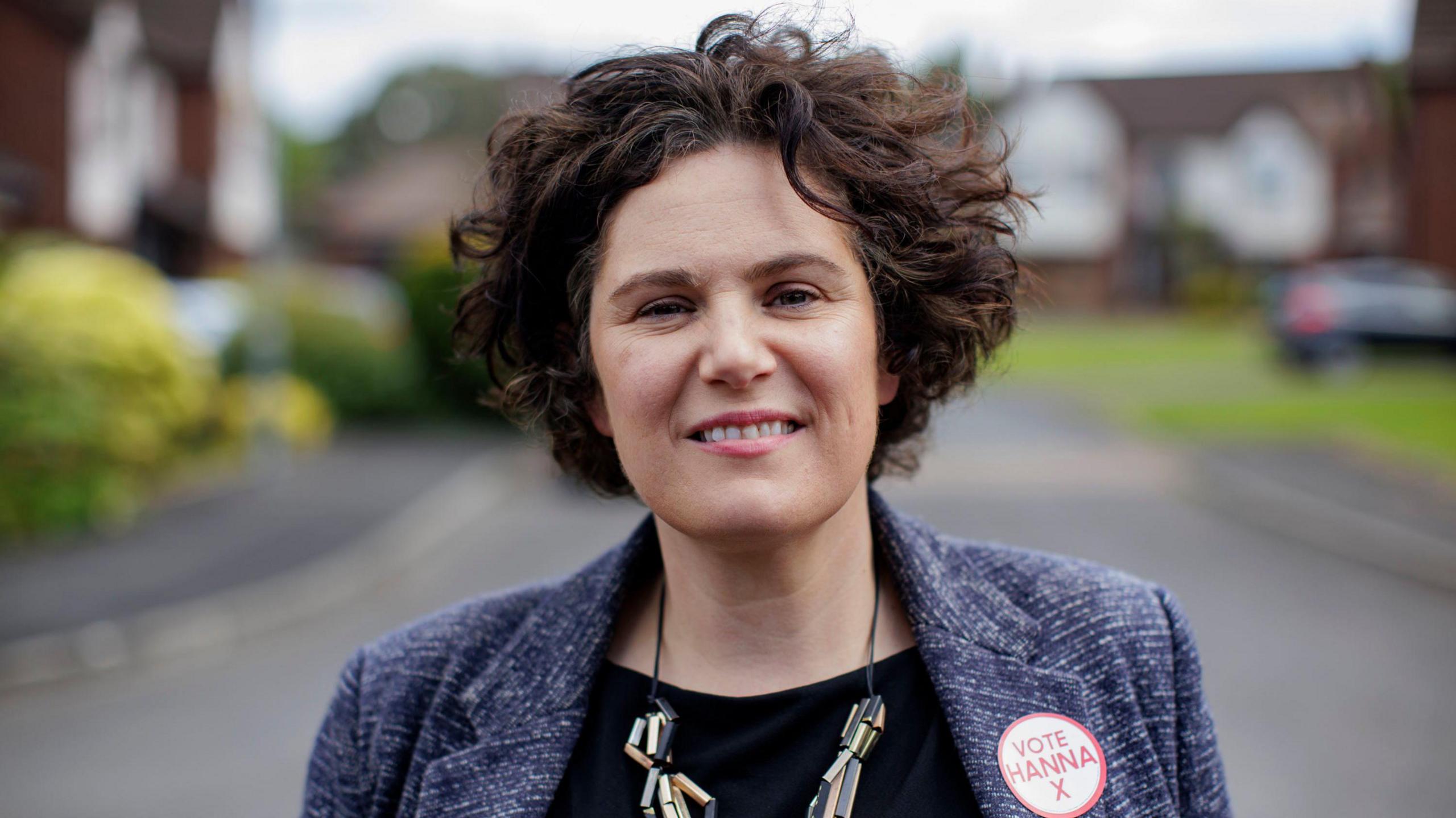 Claire Hanna smiling into the camera. A street with houses out of focus in the background. She is wearing a blazer with a sticker that reads "vote Hanna" on the lapel.