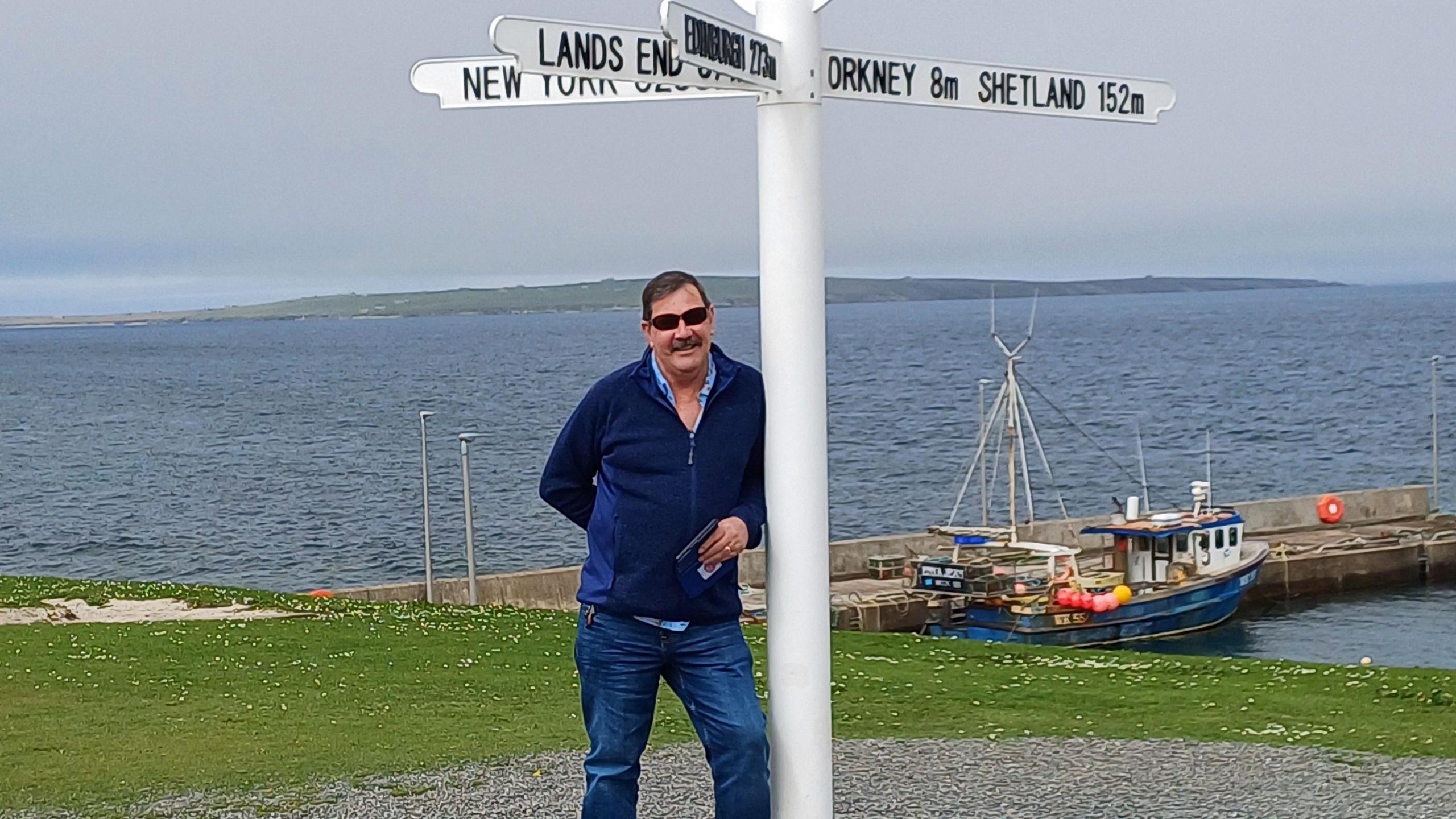 John Canning leaning on a white pole with signs pointing towards Lands End, Shetland and New York.