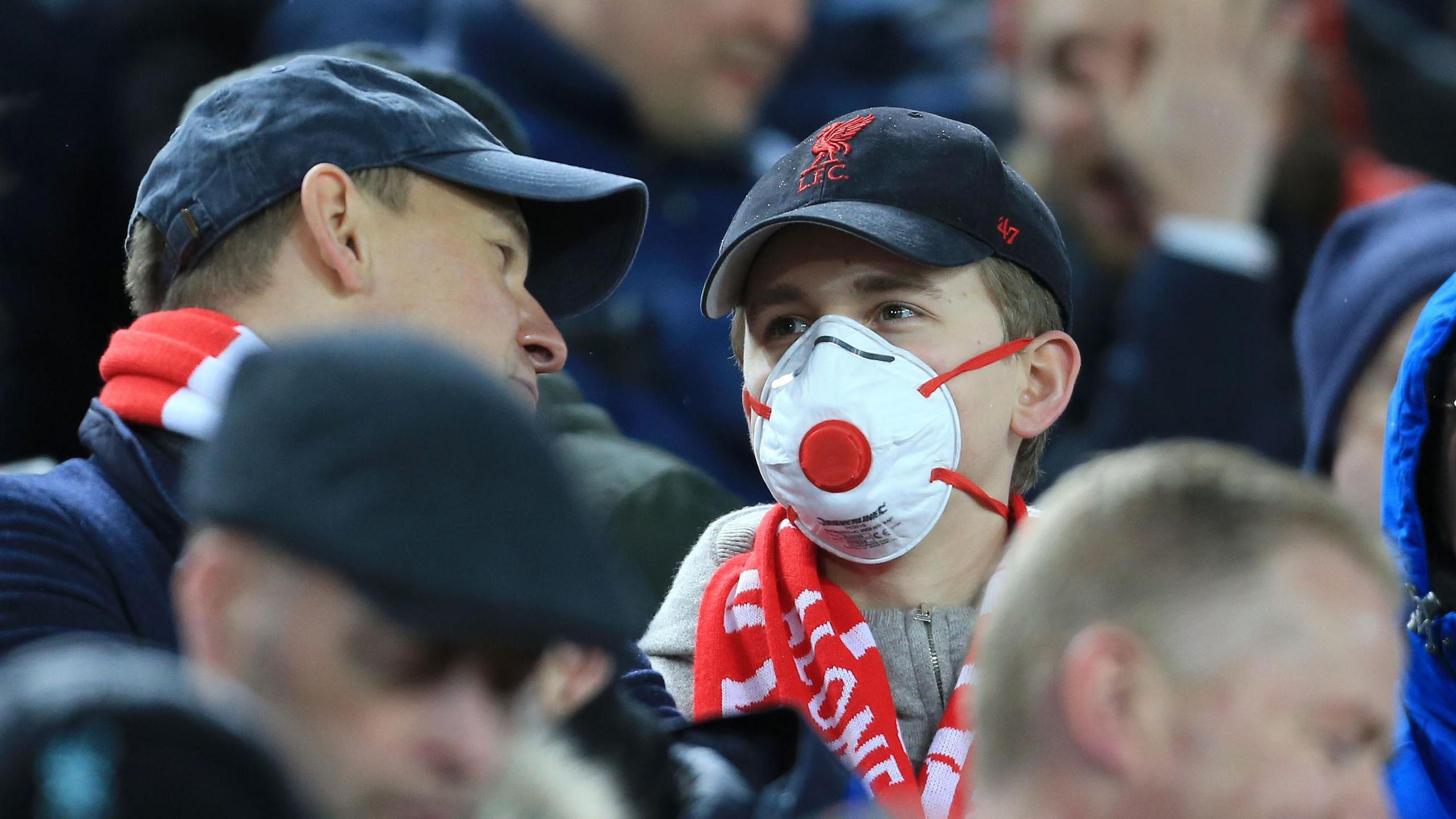 A young Liverpool fan wears a strap-on mask over his mouth and nose in the stadium, surrounded by other fans