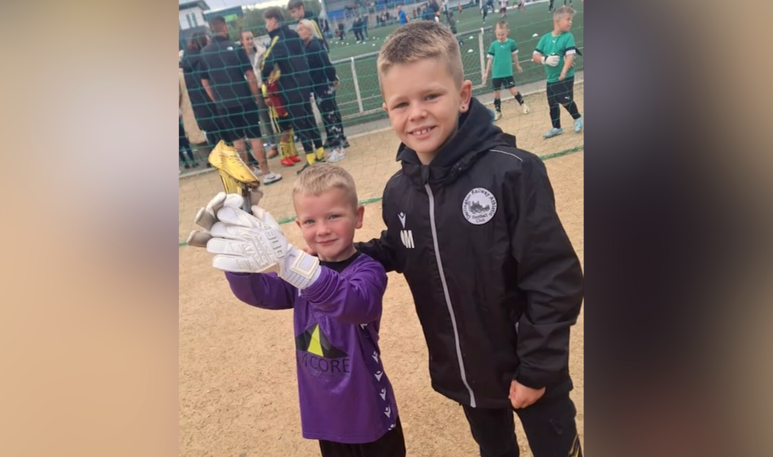 A young boy wearing a purple football shirt and white gloves holds up a gold coloured trophy while standing next to his brother, who is wearing a black Darlington Railway Athletic branded jacket. 