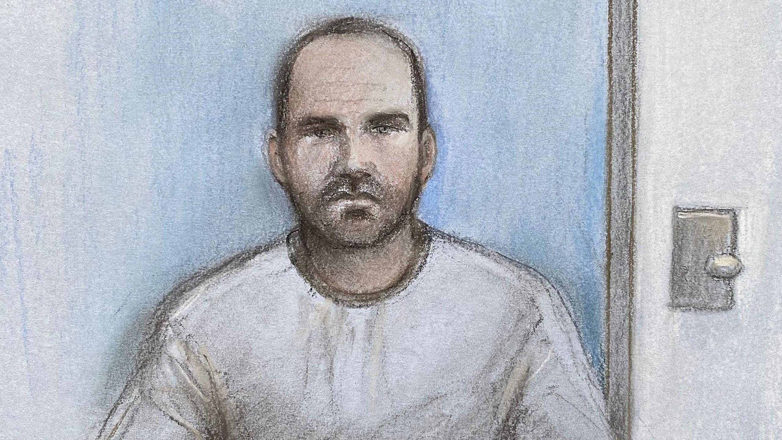 A court artist sketch of Julian Mead. He is wearing a prison-issue grey sweatshirt, has short dark hair and a short dark beard. Behind him is a blue wall and a door on the right. 