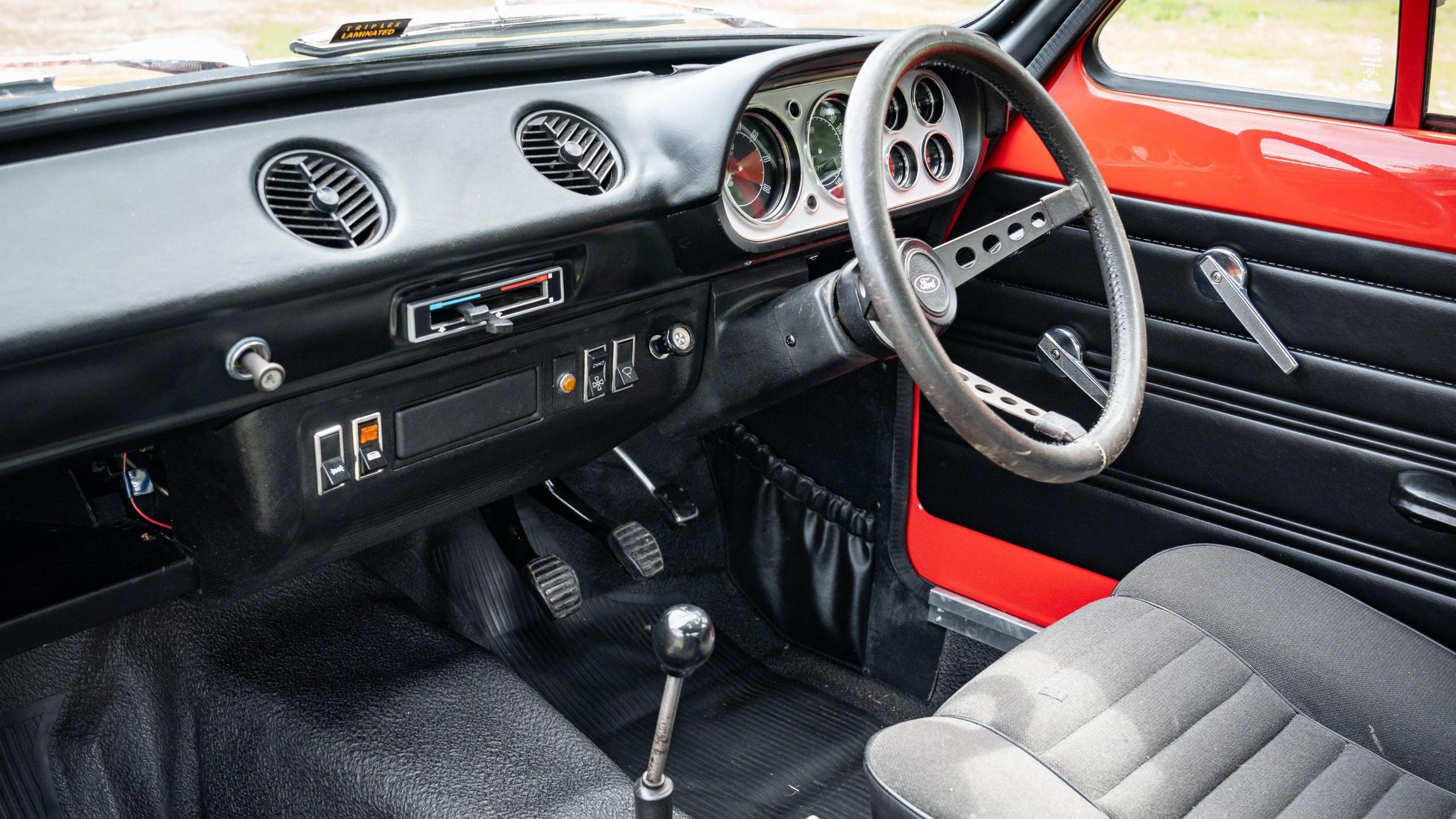 Inside the car, where there is a vintage looking steering wheel, dashboard, gear stick, radio and air-con vents.