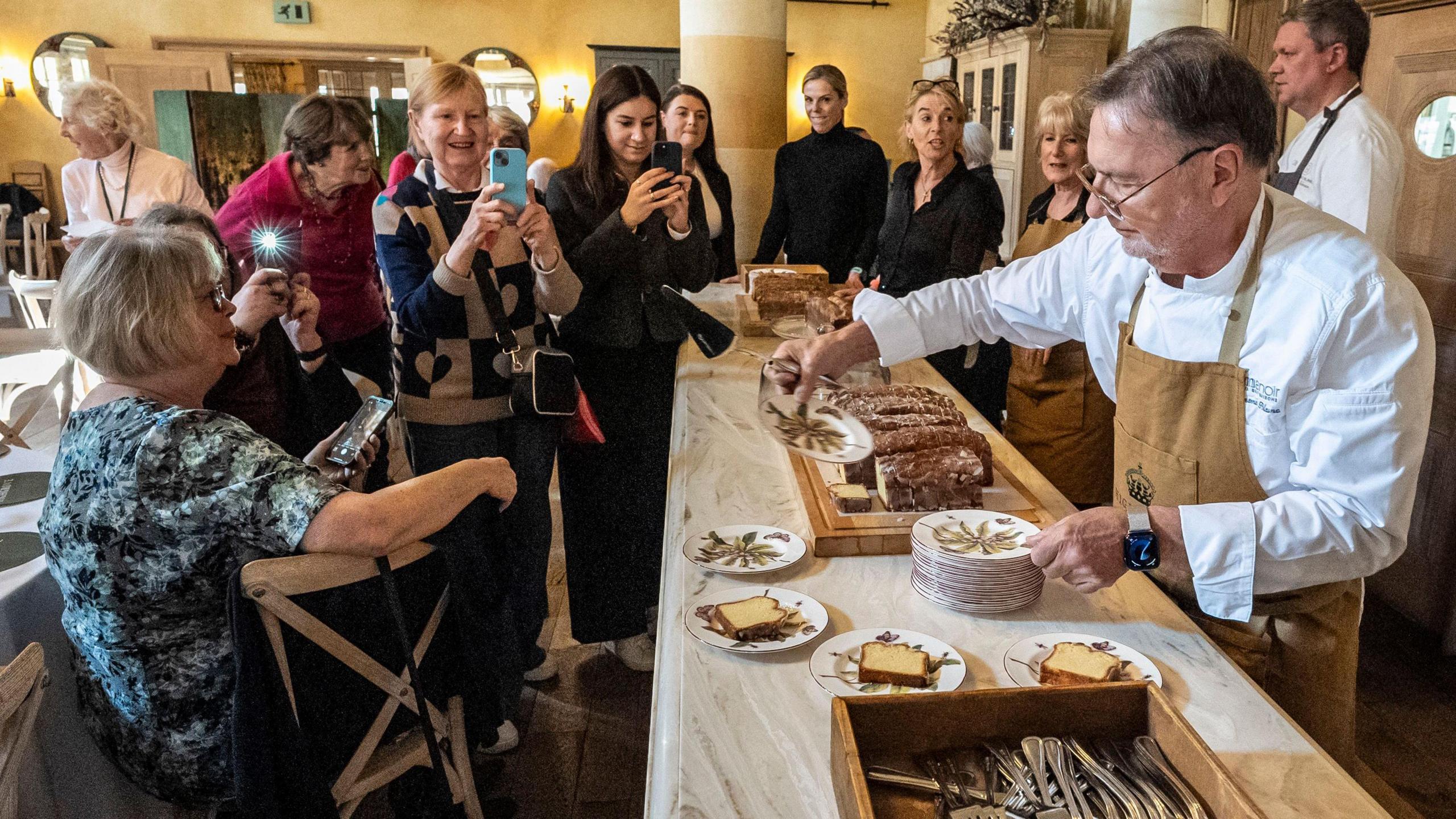 Celebrity chef Raymond Blanc serving up some food to a group of people in a large room at Highgrove. Several of the people watching are taking photos on their phones.