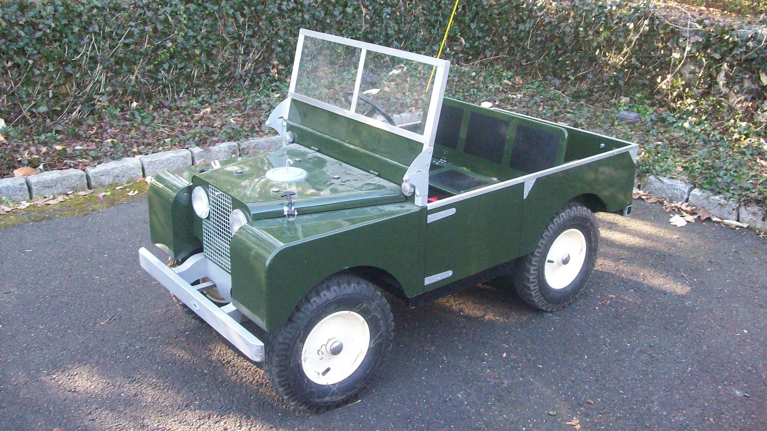 A child-sized hand-made mini-Land Rover. It is green and parked on a quiet road.