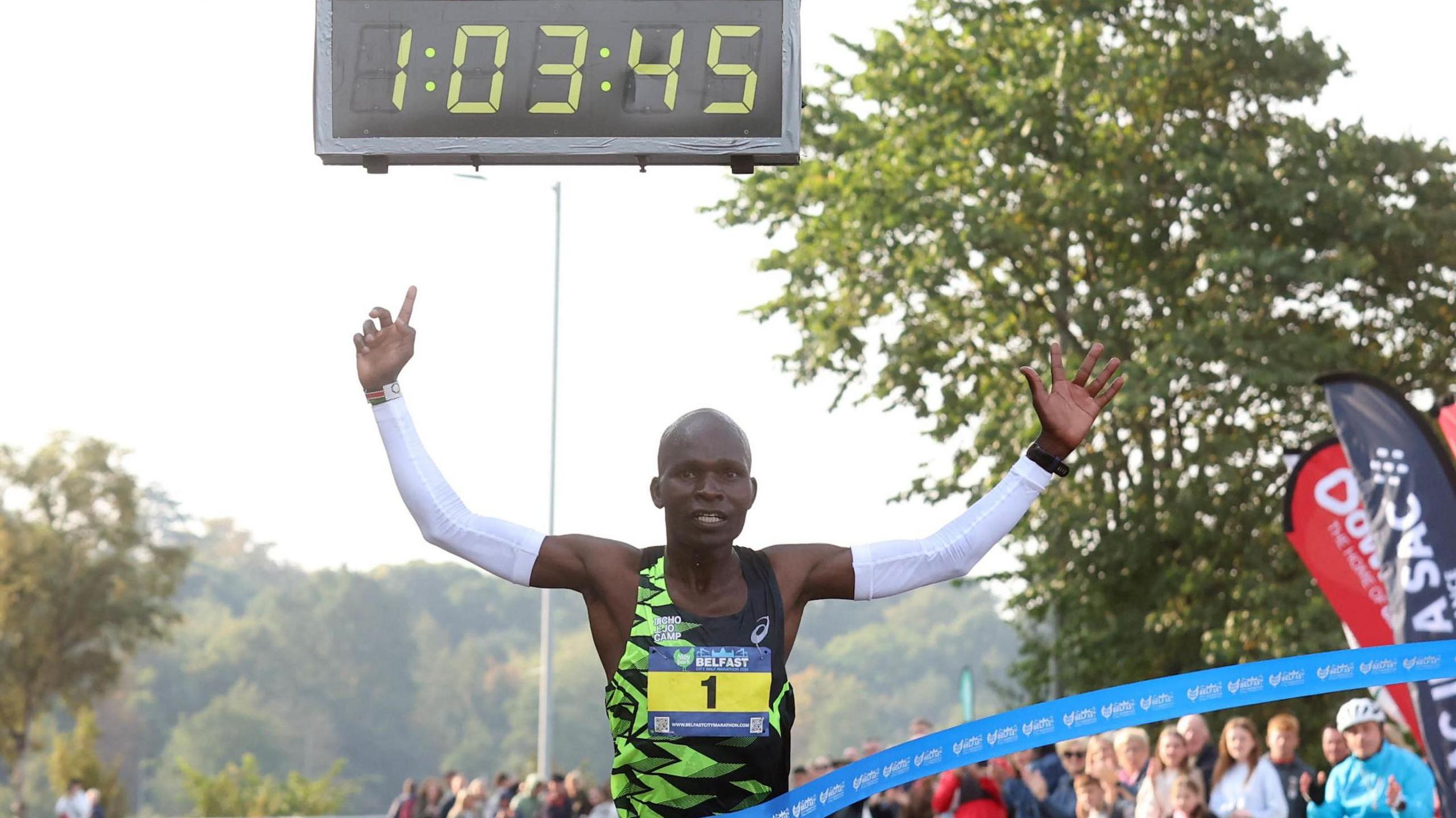 A man with his arms in the air as he crosses the finish line. The digital clock above him reads 1:03:45 as spectators look on