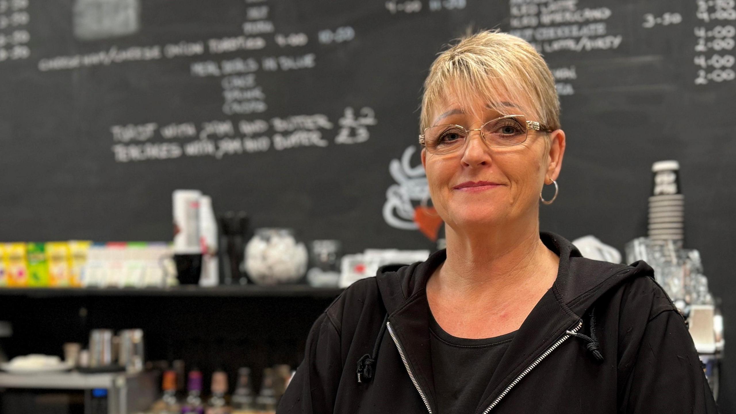 Anne Britt stands at the counter of her cafe. She has short blonde hair and wears glasses, gold-ring earrings, a black T-shirt and a black hoodie. Behind her, the menu is written in white chalk on a large blackboard.