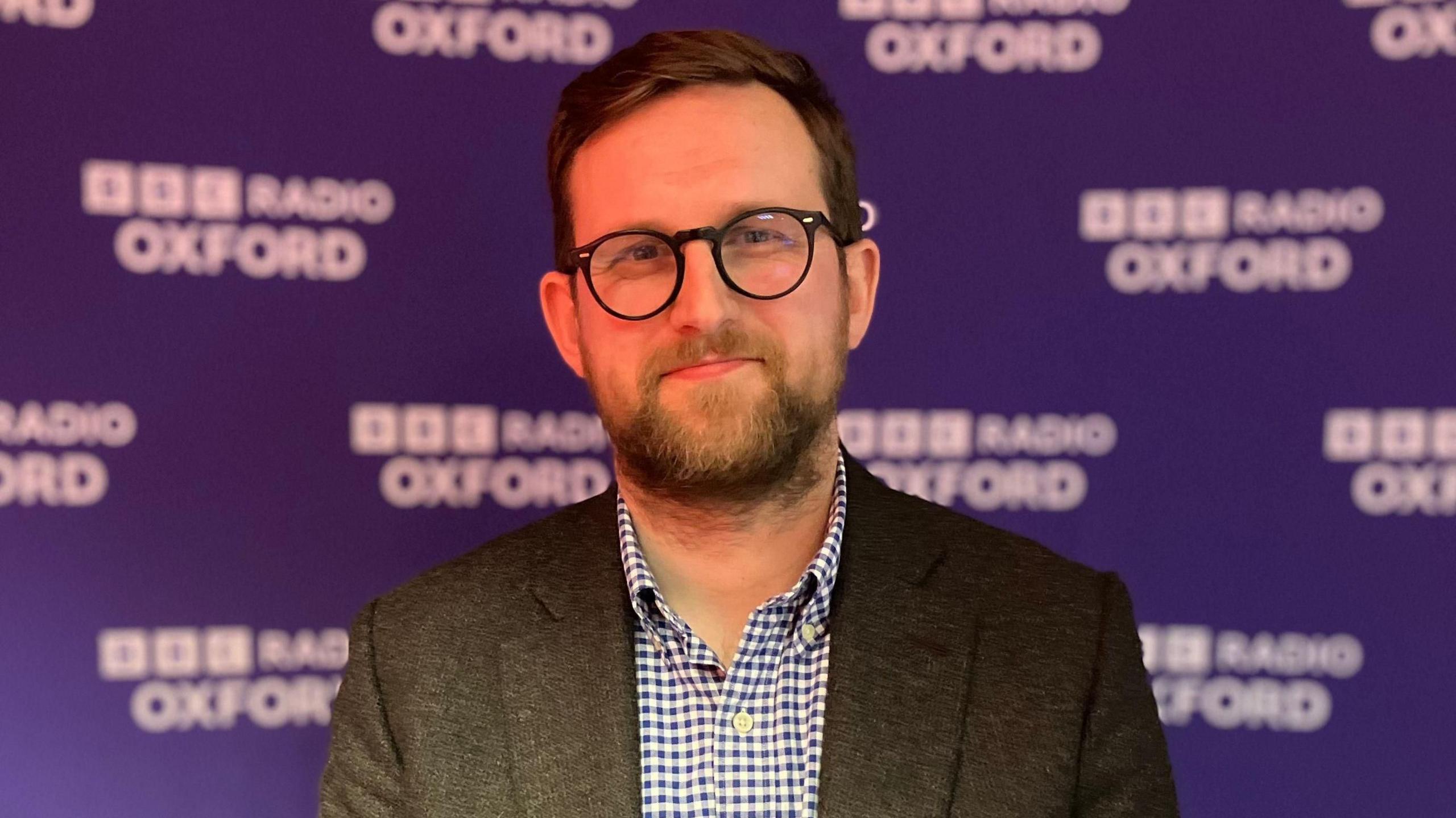 Freddie van Mierlo stands in front of a purple banner with BBC Radio Oxford branding. He has short dark hair, black rimmed glasses, and wears a dark suit jacket with a checked shirt.