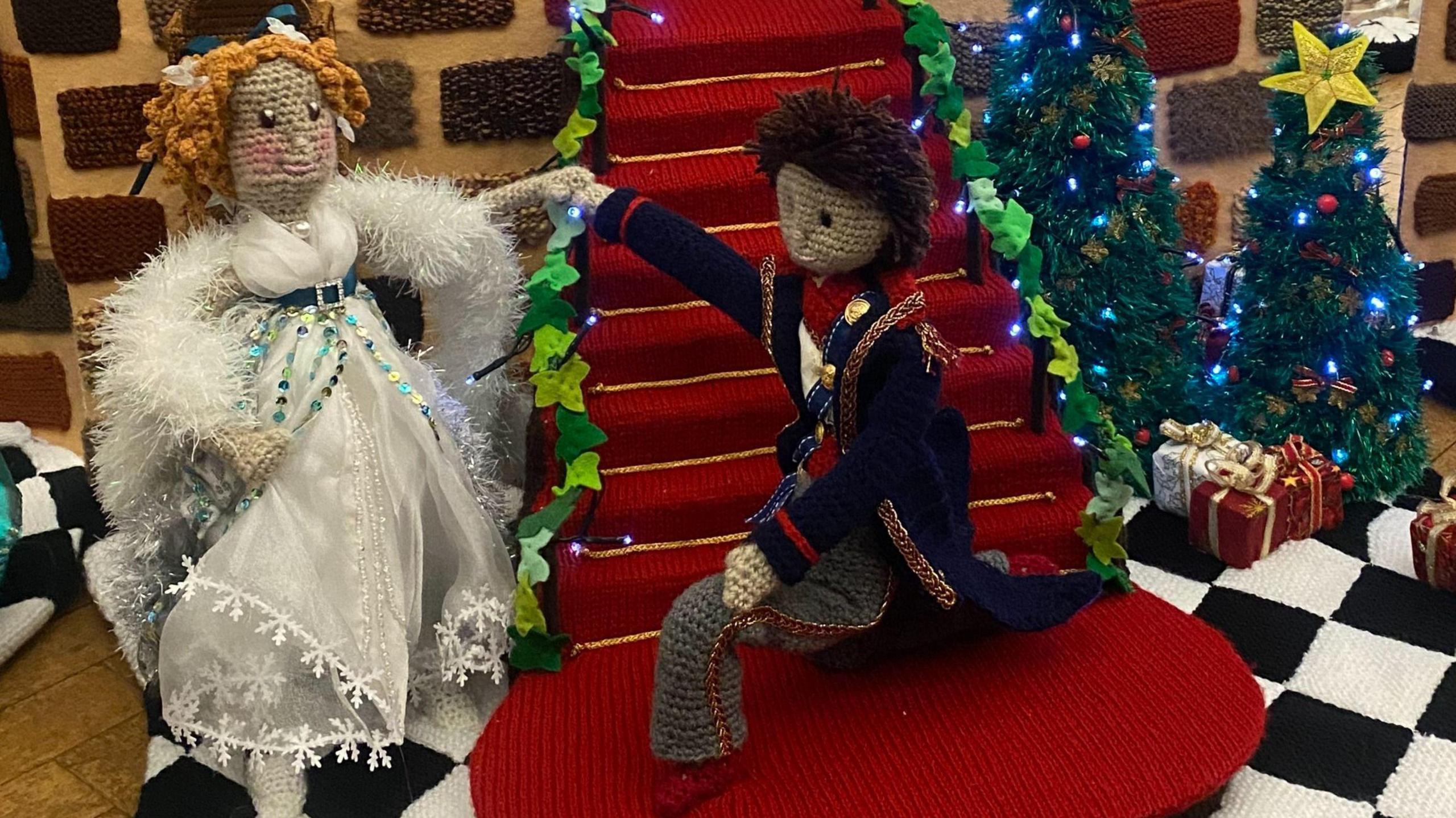 Cinderella and Prince Charming knitted, showing Cinderella in a white dress and ringlets in her hair. She is standing by a red carpet on stairs, next to Prince Charming, who is kneeling down. There are two Christmas trees to the side. 